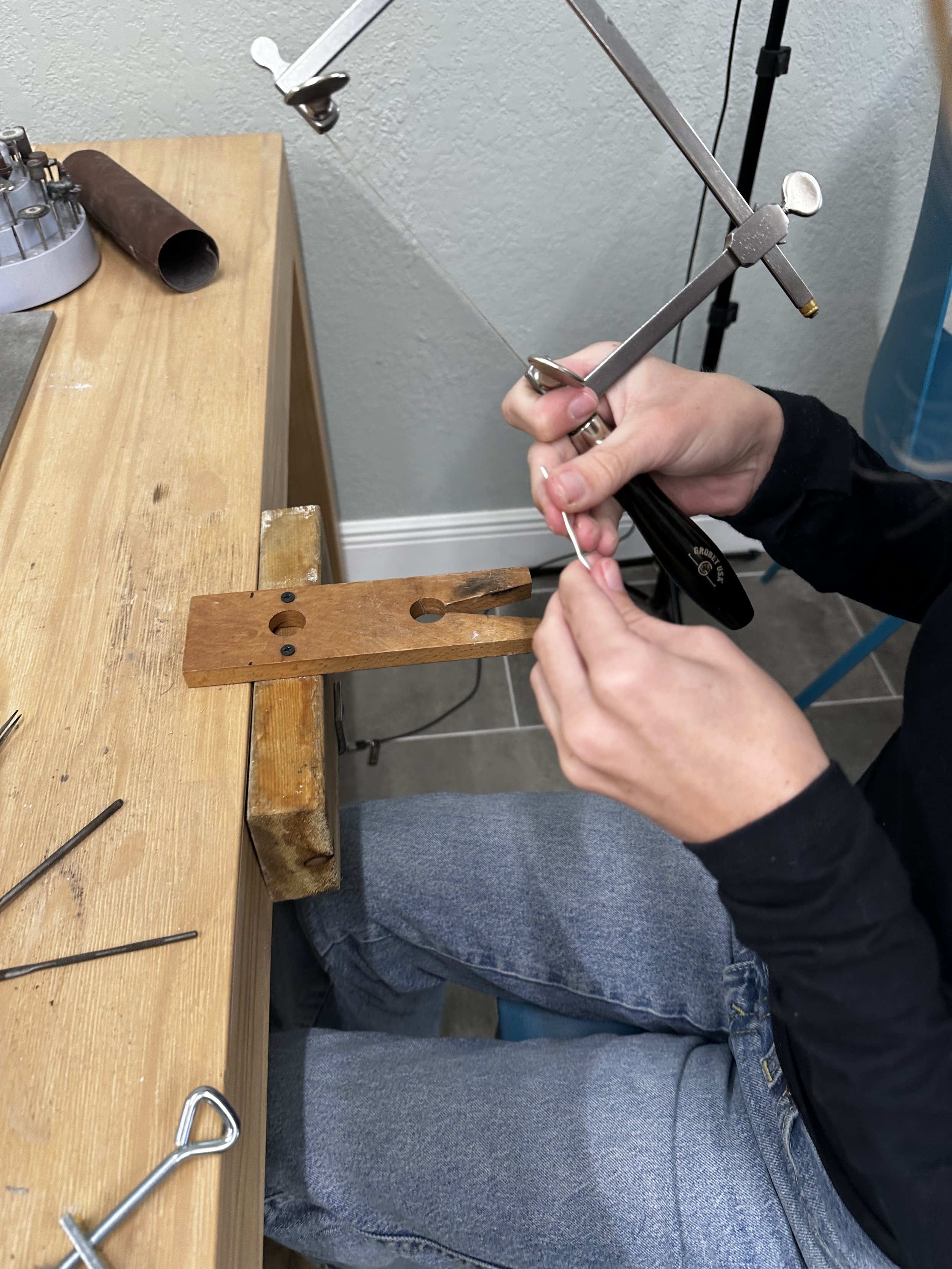 Person working on a craft project using a coping saw at a wooden workbench with various tools, including a metal rod and a wooden block, in an indoor setting.