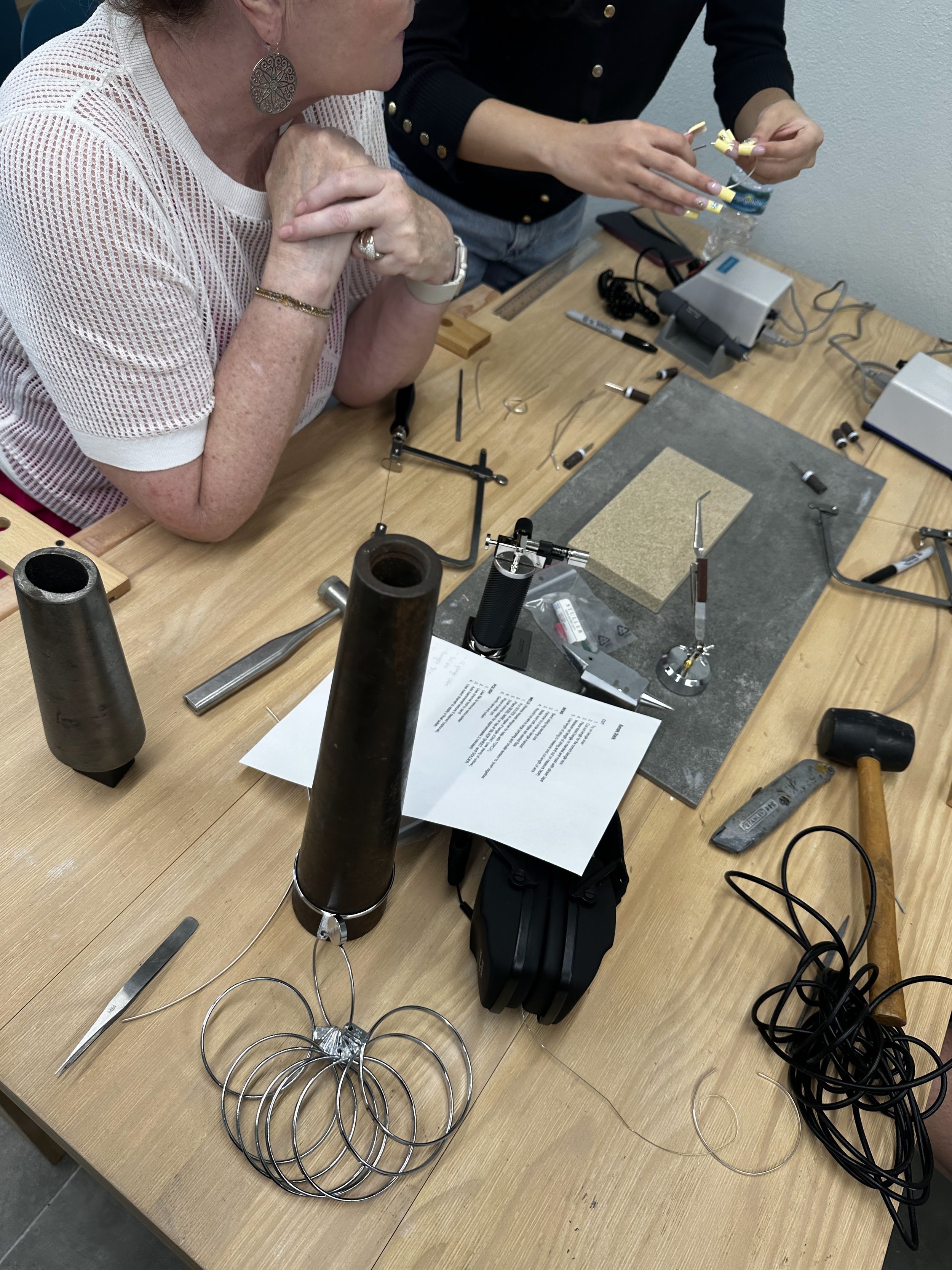 Two women working on jewelry making at a workshop table, with tools, jewelry components, and papers scattered on the surface.