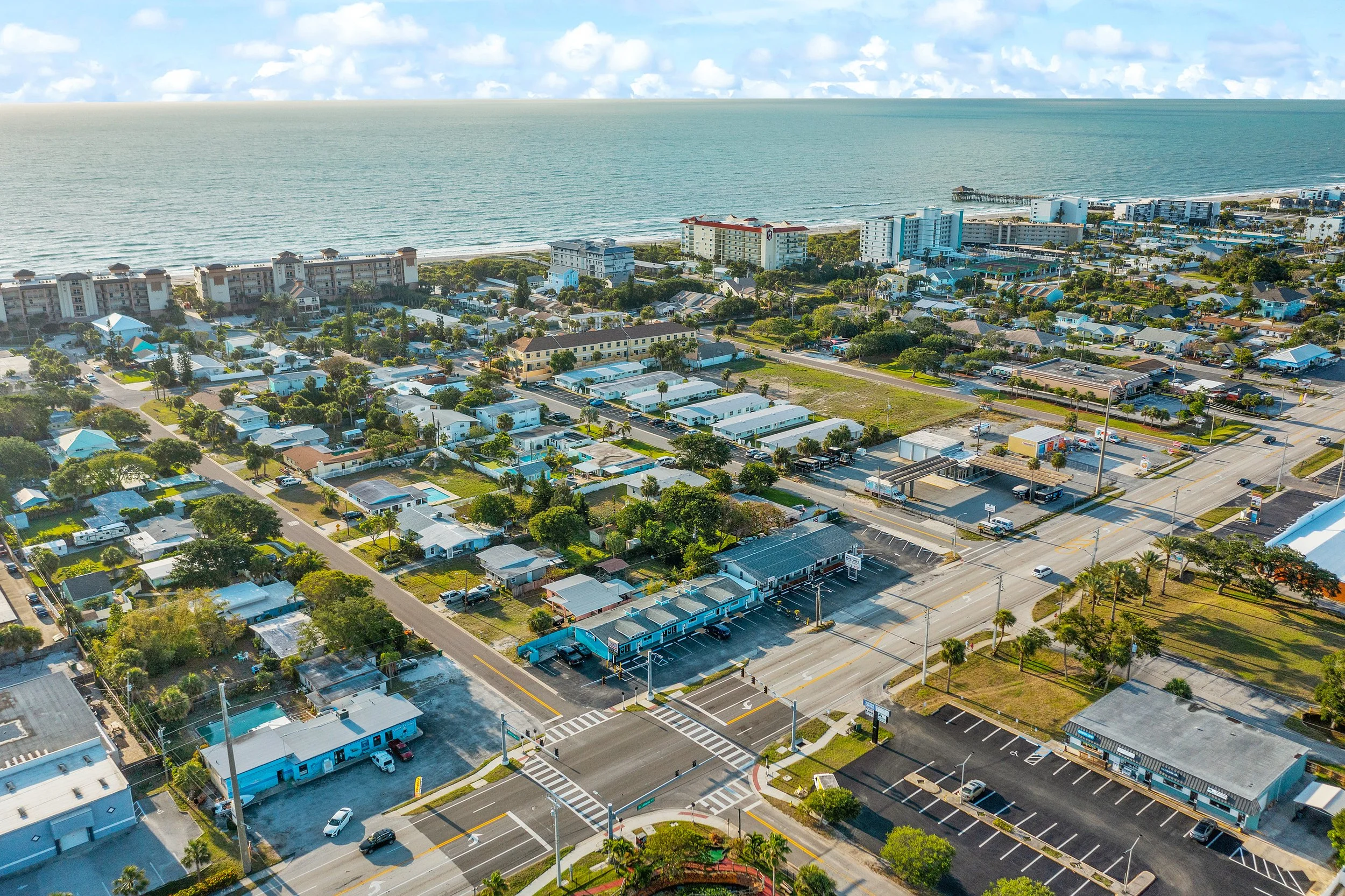 Aerial view of a coastal city with residential buildings, commercial establishments, parking lots, and a beach with ocean in the background. Clear sky with some clouds.