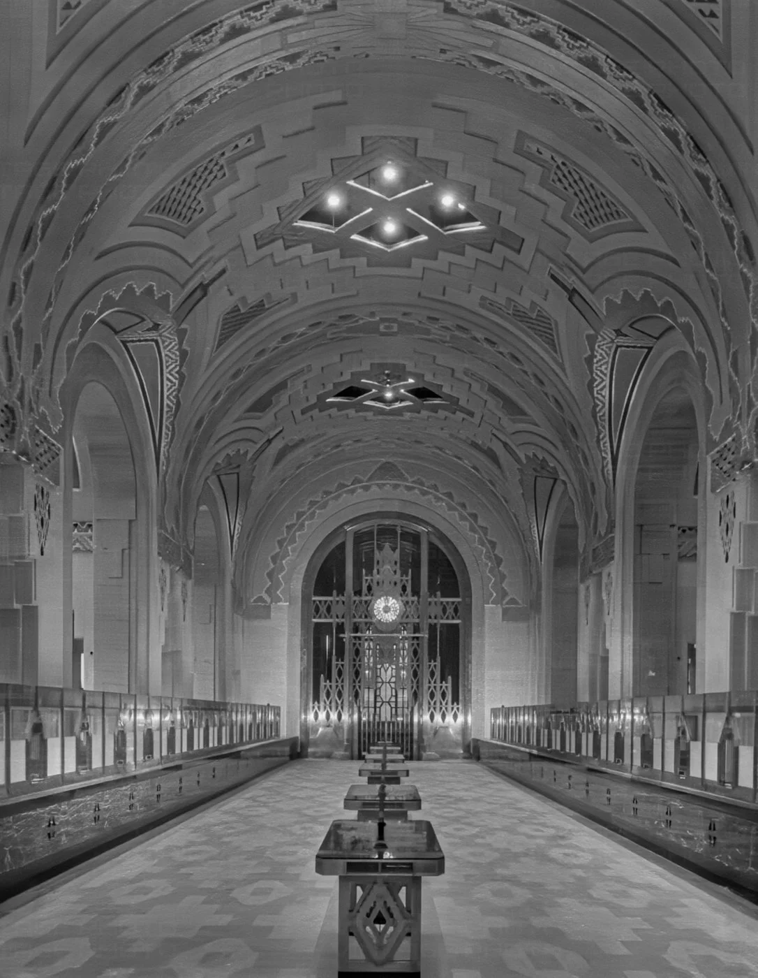 Interior of a grand, ornate building with high, arched ceilings, intricate ceiling decorations, and a large clock on the far wall, illuminated by chandeliers.