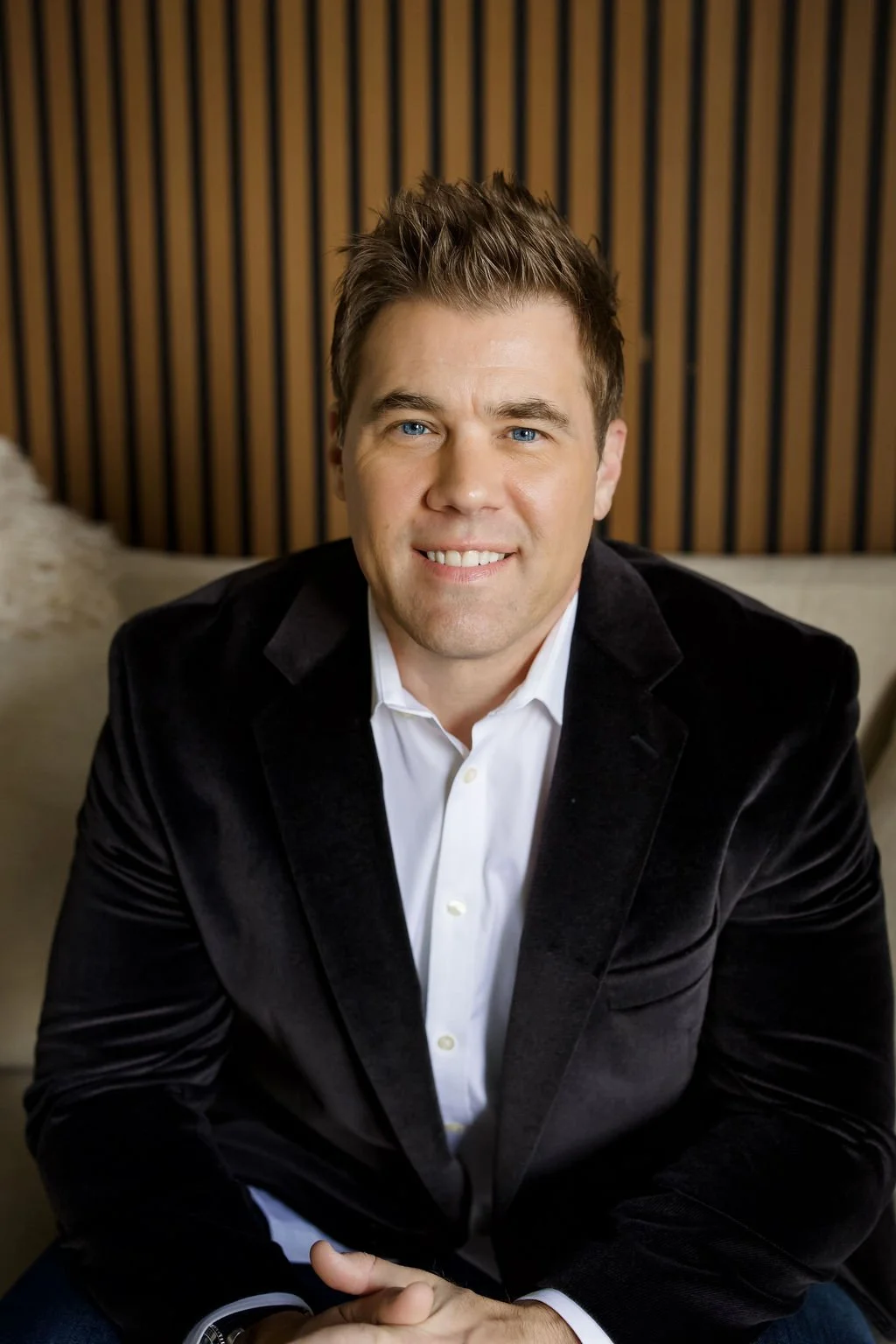 Portrait of a smiling man with blue eyes, brown hair, wearing a black jacket and white shirt, sitting indoors against a wooden slatted wall.