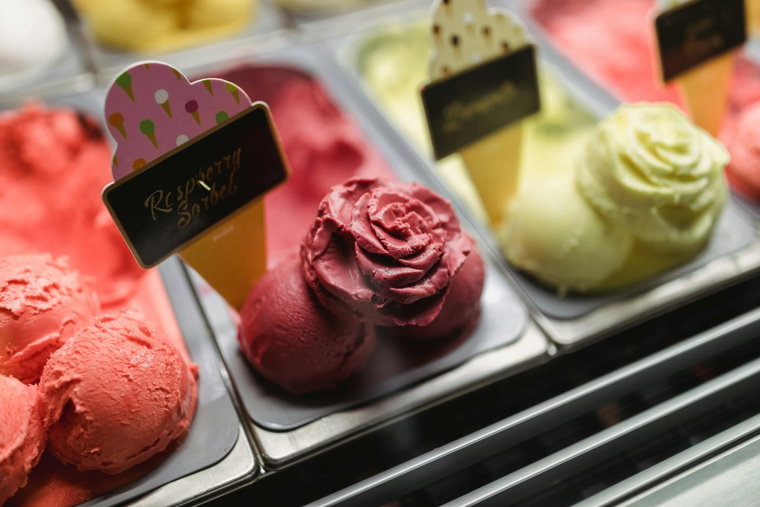 Close-up of colorful gelato scoops in a display case, including pink, red, and yellow flavors with decorative signs.
