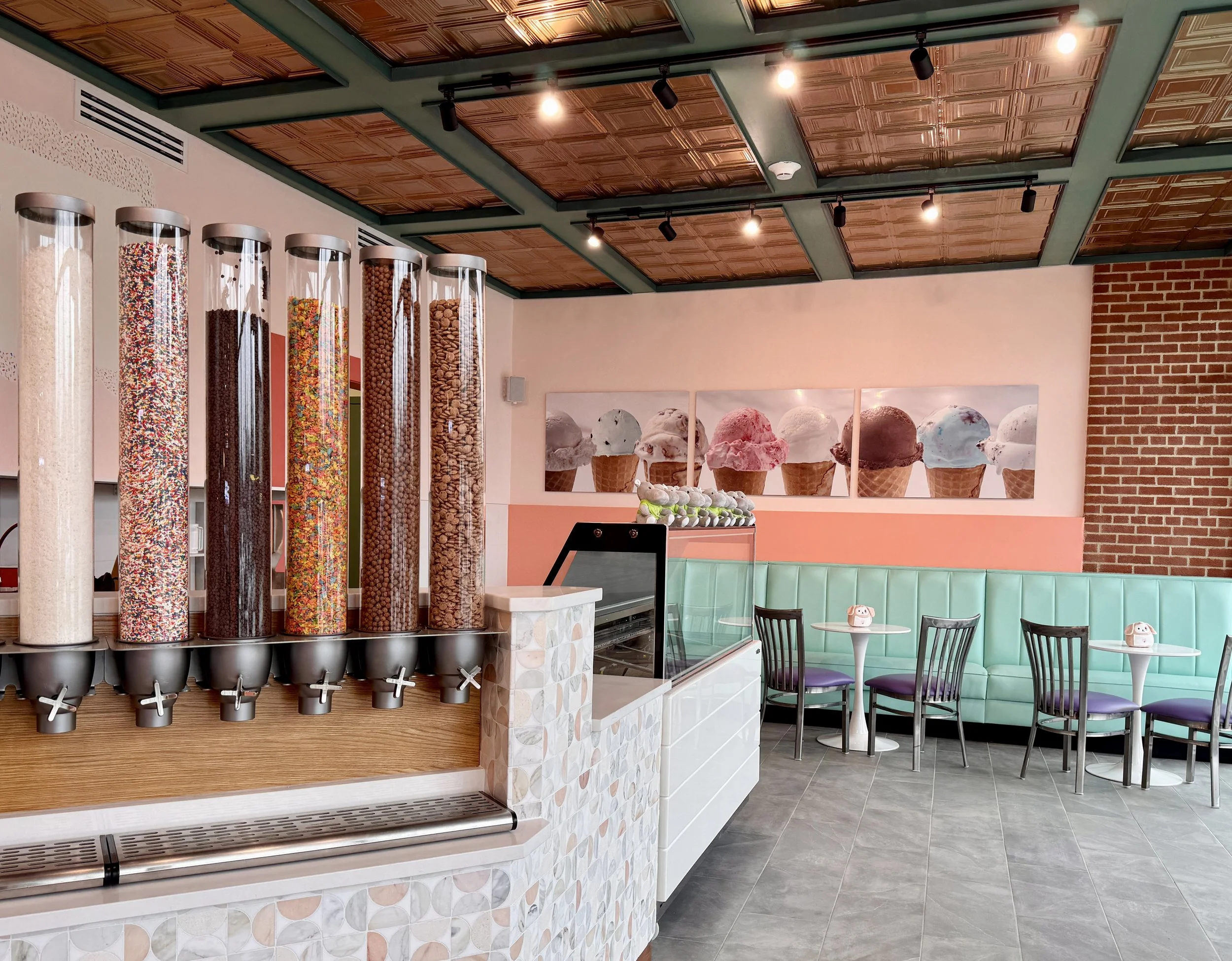 Interior of the Gelati Biscoti store, showcasing a row of topping dispensers, a mint green cushioned booth with tables and chairs, a wall with gelato cone artwork, and a brick accent wall.