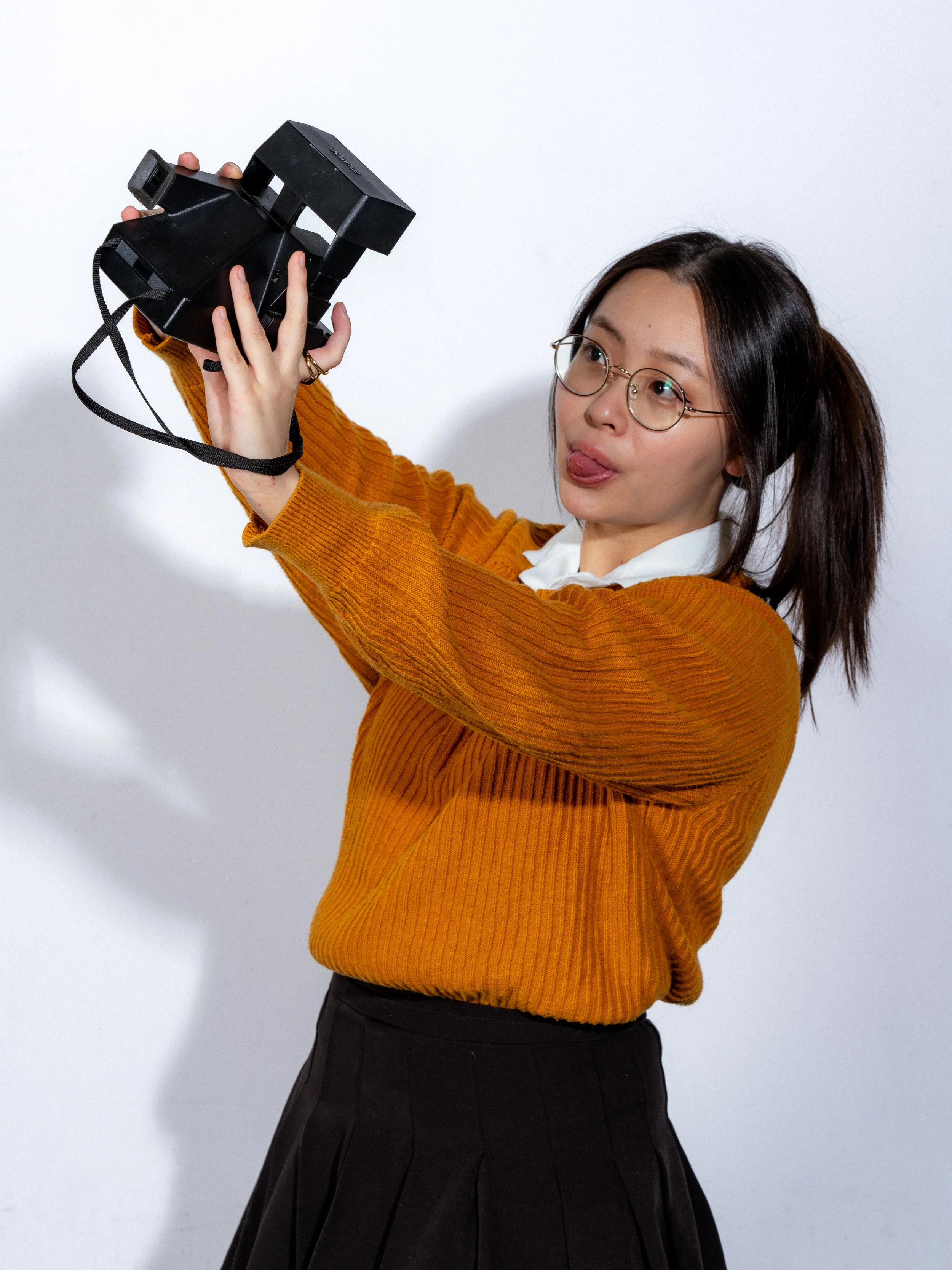 Woman with glasses taking a selfie with a vintage camera, sticking her tongue out.