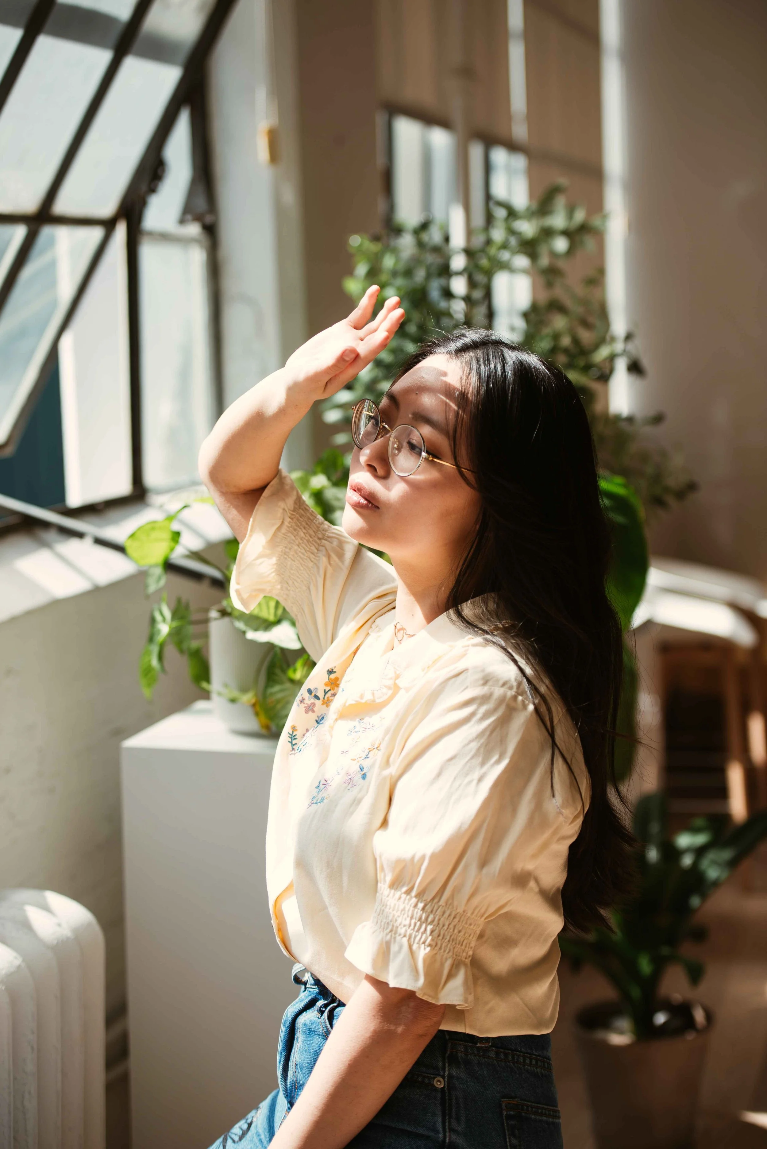 A woman with glasses and long dark hair standing near a window with sunlight, raising her hand to her forehead, surrounded by green plants in an indoor setting.