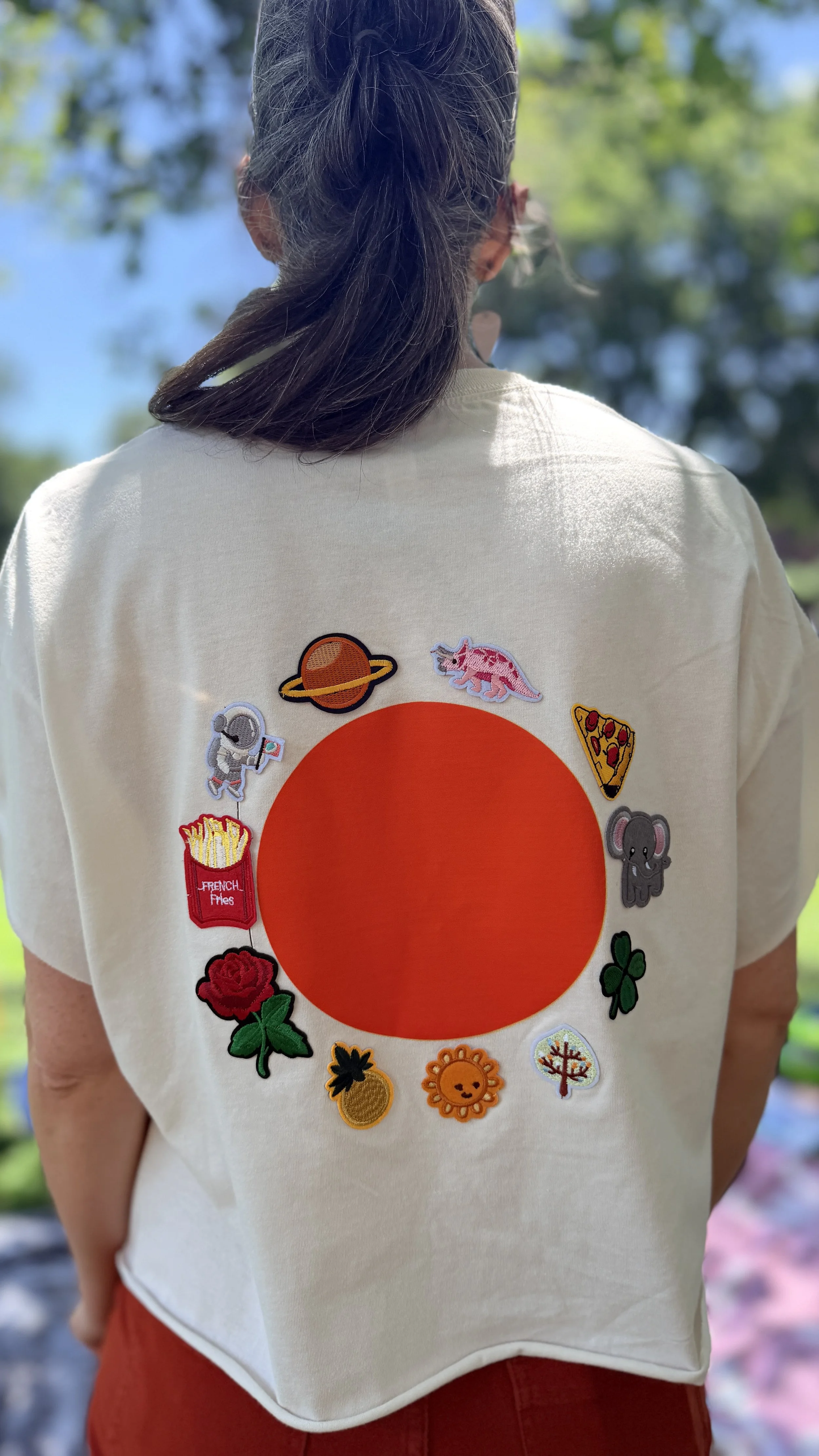 The 001 shirt. Orange dot on cream, with iron-on patches arranged in a circle around it — each one chosen by the person wearing it. Saturn, dinosaur, pizza, elephant, clover, sun, tree, pineapple, rose, fries, astronaut.