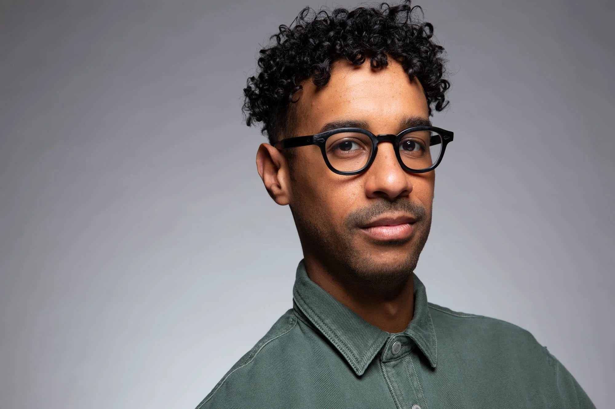 A young man with curly black hair wearing black framed glasses and a dark green shirt with a collar, standing against a plain gray background.