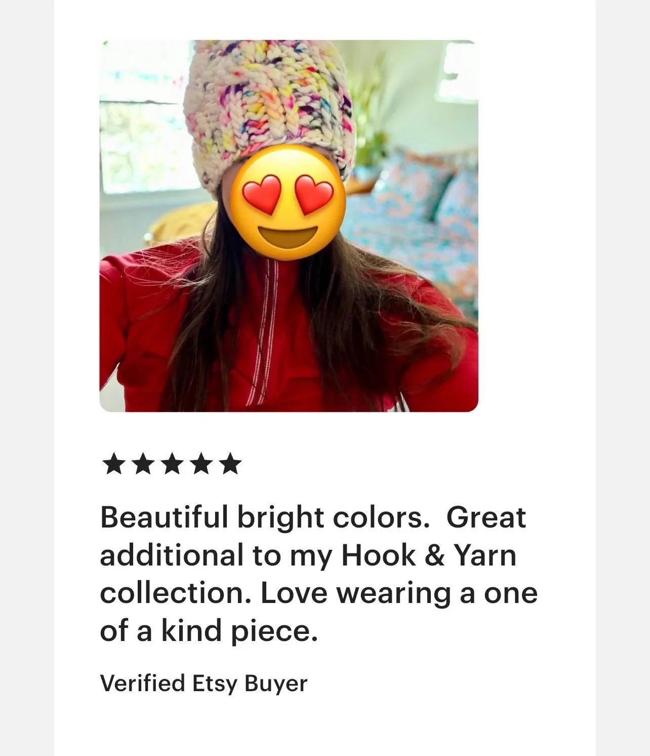 A woman wearing a colorful knit hat and a red jacket, with an emoji covering her face featuring heart eyes and a smiling face, standing in a bright room with a bed and window in the background.