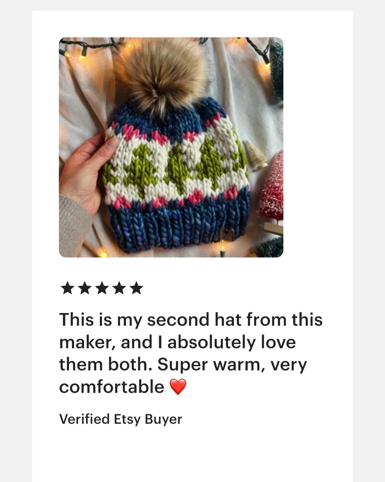 Colorful knitted winter hat with a pom-pom on top, featuring green reindeer patterns, being held by a person. Christmas lights and a red ornament are in the background.