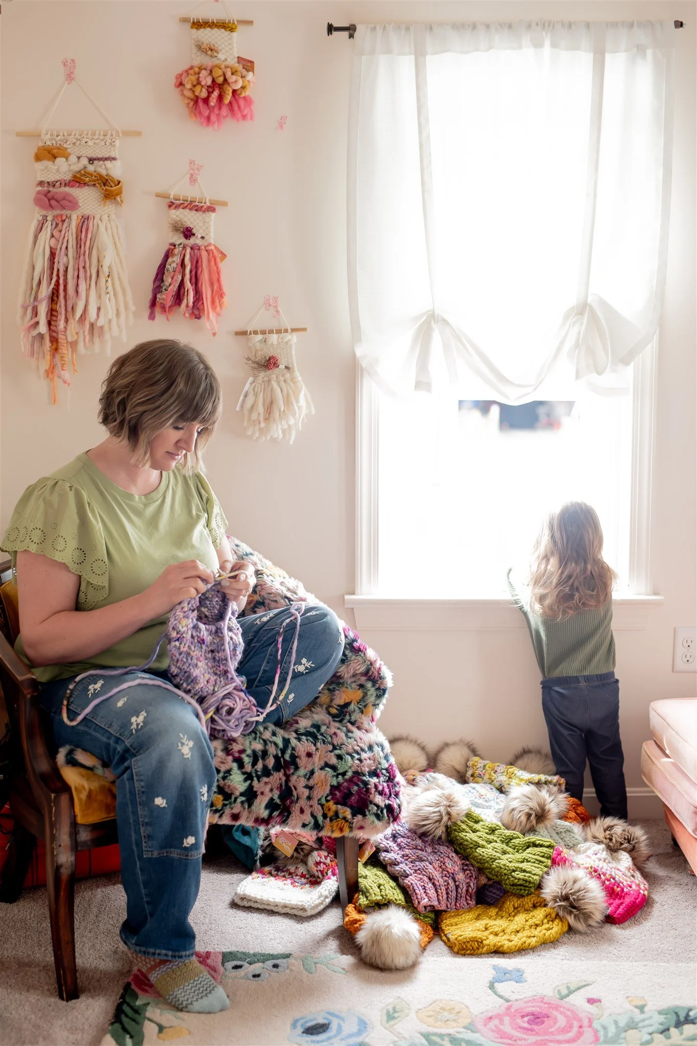 A woman knitting while seated on a chair in a sunlit room, with a young girl looking out of a window. The wall behind them is decorated with colorful woven wall hangings. There are piles of colorful knitted hats on the floor.
