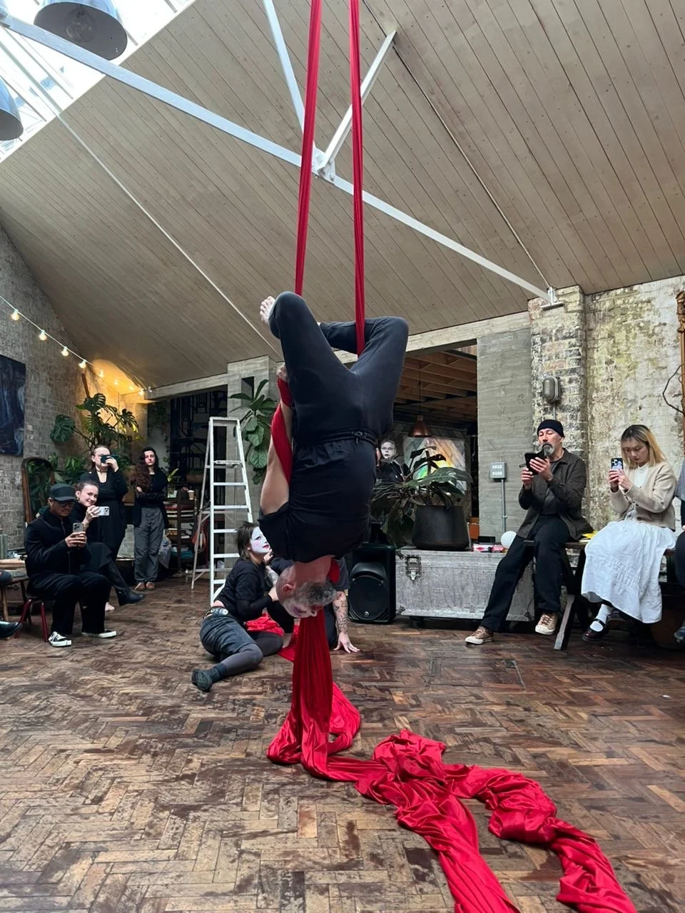 An aerial performer hangs upside down from long red silks that spill across the wooden floor, as a close audience watches intently in an industrial, site-specific performance space.