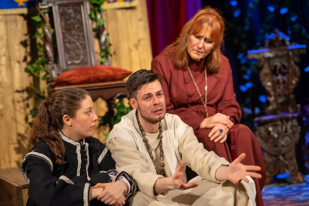 Richard II sits distressed and open-handed beside Aumerle, while the Bishop of Carlisle watches gravely from behind, in a richly dressed court scene from Richard II by William Shakespeare.