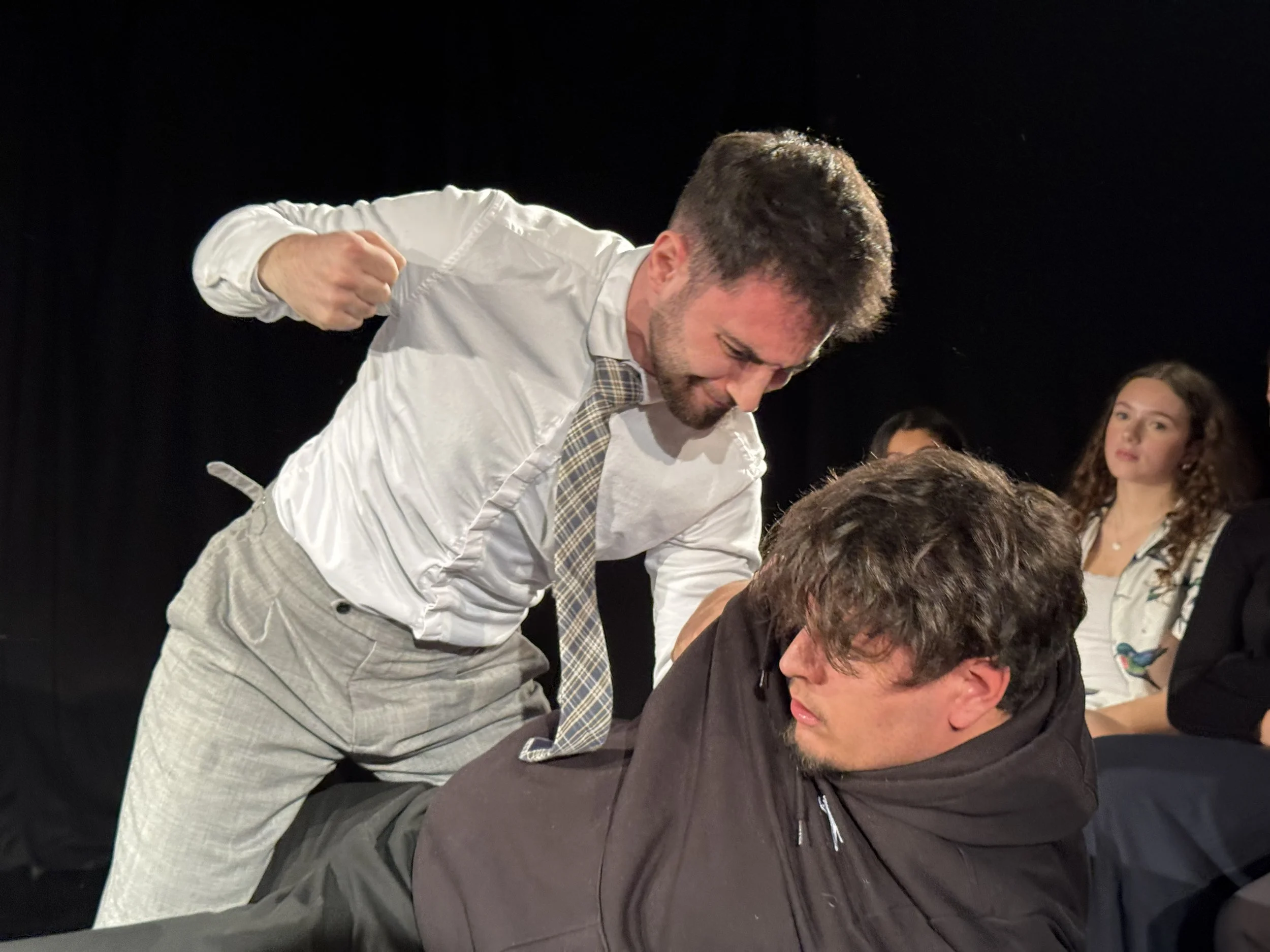 Tom, the agency tutor, looms over Daryll, a student in a hoodie, his fist clenched as if to strike while gripping his shoulder under stark stage lighting in a tense scene from Monster by Duncan Macmillan.