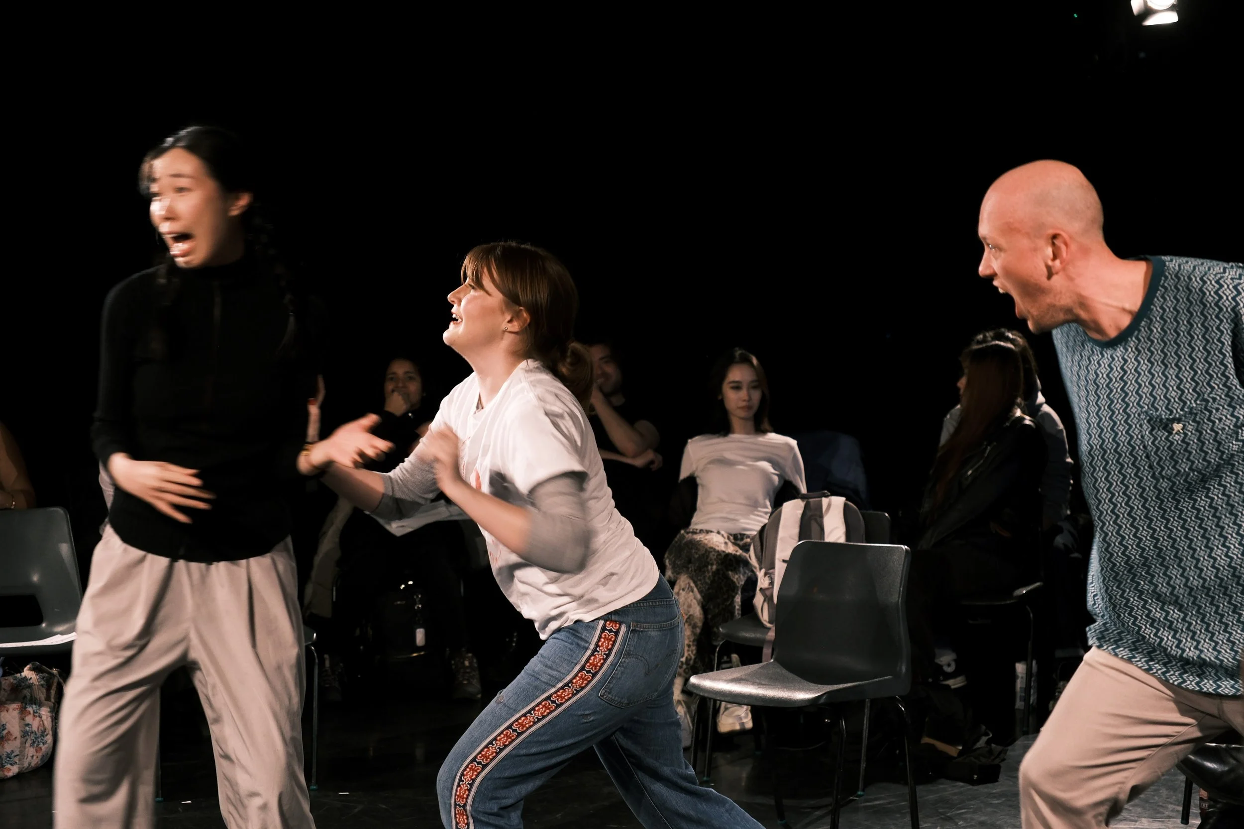 Three actors perform an improvised scene in the round, reacting with heightened emotion under stark lighting as an audience watches closely from surrounding chairs, creating an immediate and dynamic performance atmosphere.
