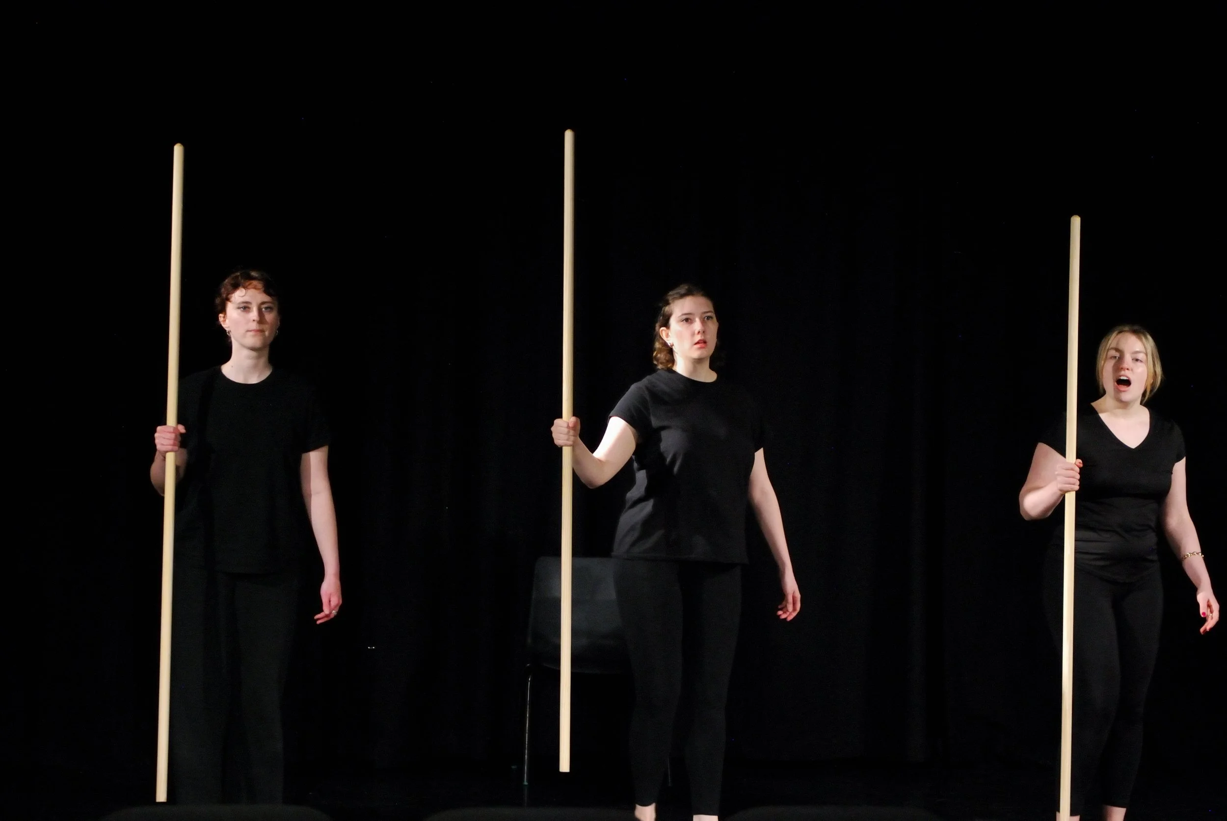 Three performers in black hold wooden staffs during Weyward Sisters, a physical theatre piece inspired by Macbeth and featuring Song of the Goat Theatre, with minimalist staging, ensemble movement, and ritualistic performance imagery.