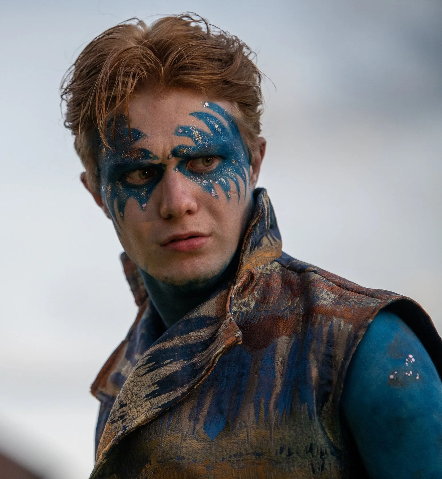 Puck, with vivid blue face paint sweeping across his eyes and body, stands in a textured, earth-toned costume against an open sky, captured in a striking outdoor performance of A Midsummer Night’s Dream by William Shakespeare.