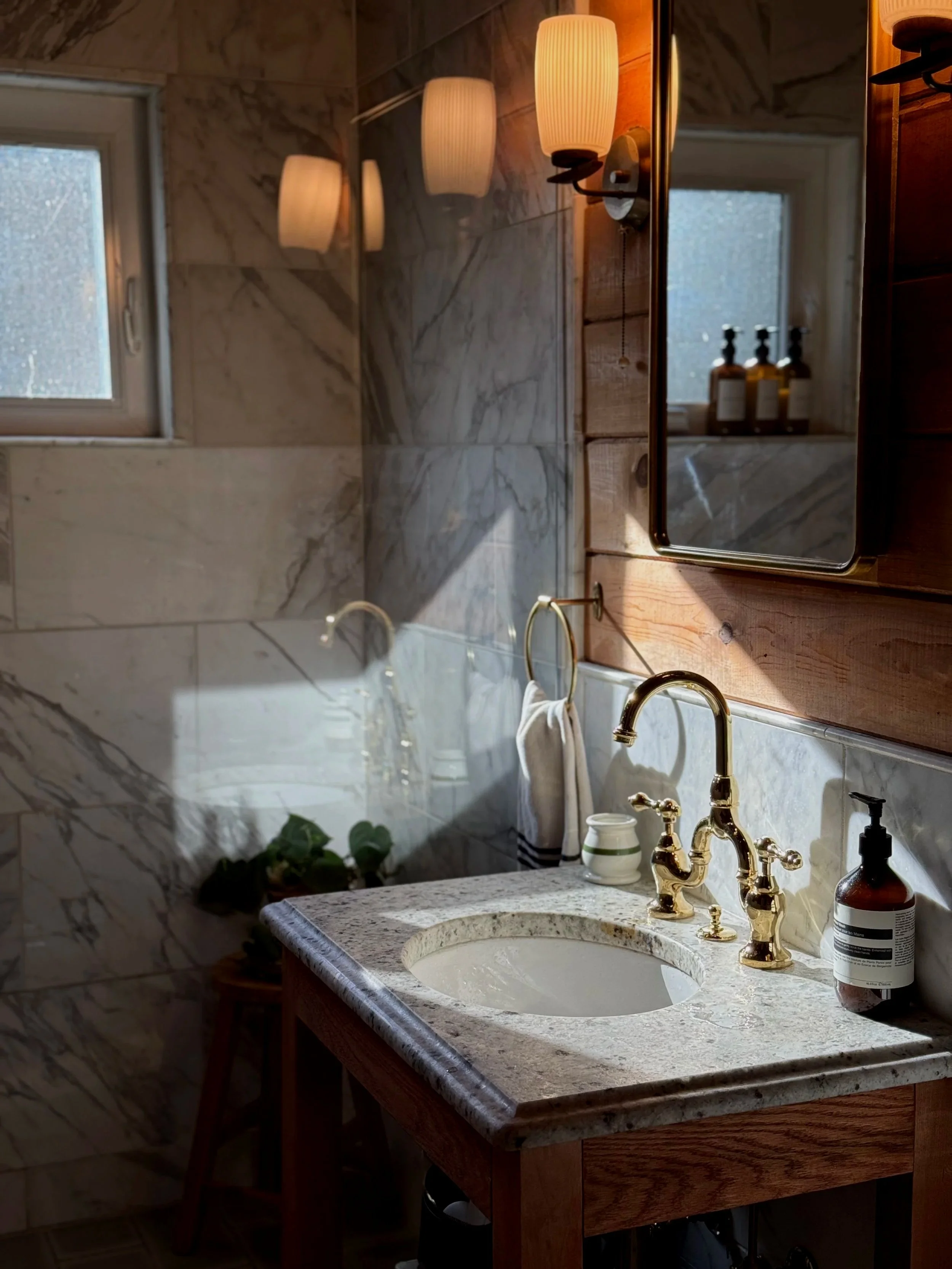 A bathroom vanity with a marble countertop, gold faucet, and a mirror reflecting a wall-mounted light. A small potted plant is on a stool near the window, and a soap dispenser is on the right side of the sink. The wall behind the vanity features wooden paneling and marble tiles, with towels hanging on hooks.