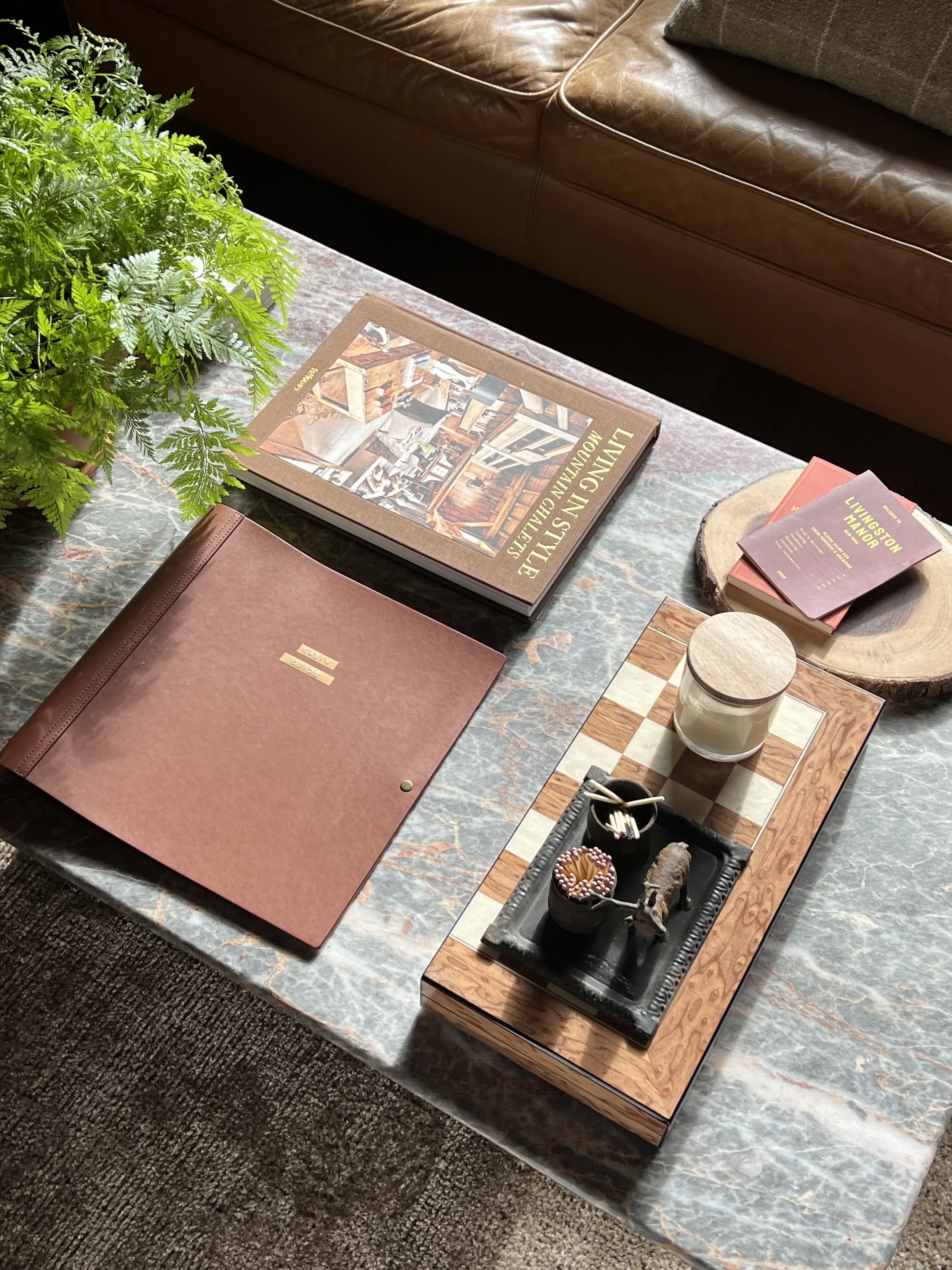 A marble table with various decorative items, including a large green fern plant, a brown leather menu, a book titled "Living in Style" showcasing interior design, a small stack of pamphlets titled "Livingston Manor," a wooden tray with a candle, small bowl of toothpicks, a bowl of candies, and a small animal figurine.