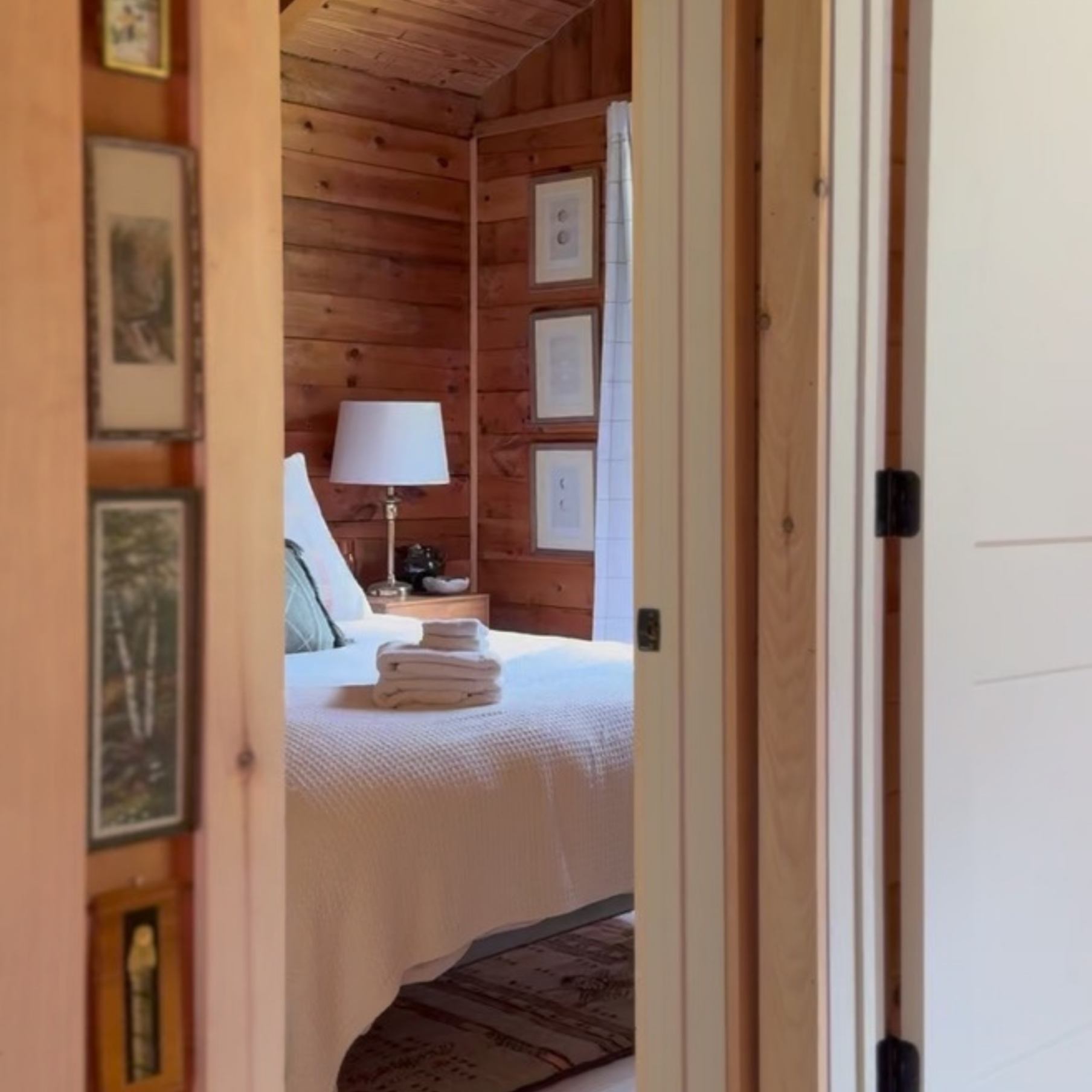 A view through a partially opened door showing a bedroom with wooden walls and ceiling, a bed with white bedding and folded towels, a bedside table with a lamp, framed pictures on the wall, and a window with curtains.