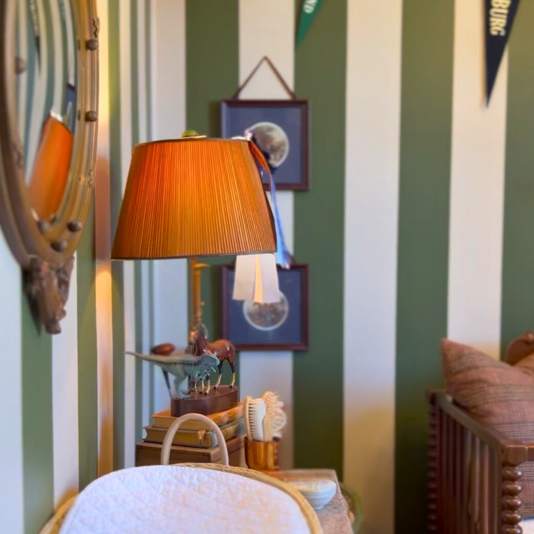 A vintage-style room with a green and white striped wall, a table lamp with an orange shade, framed pictures, and a sofa with pillows.