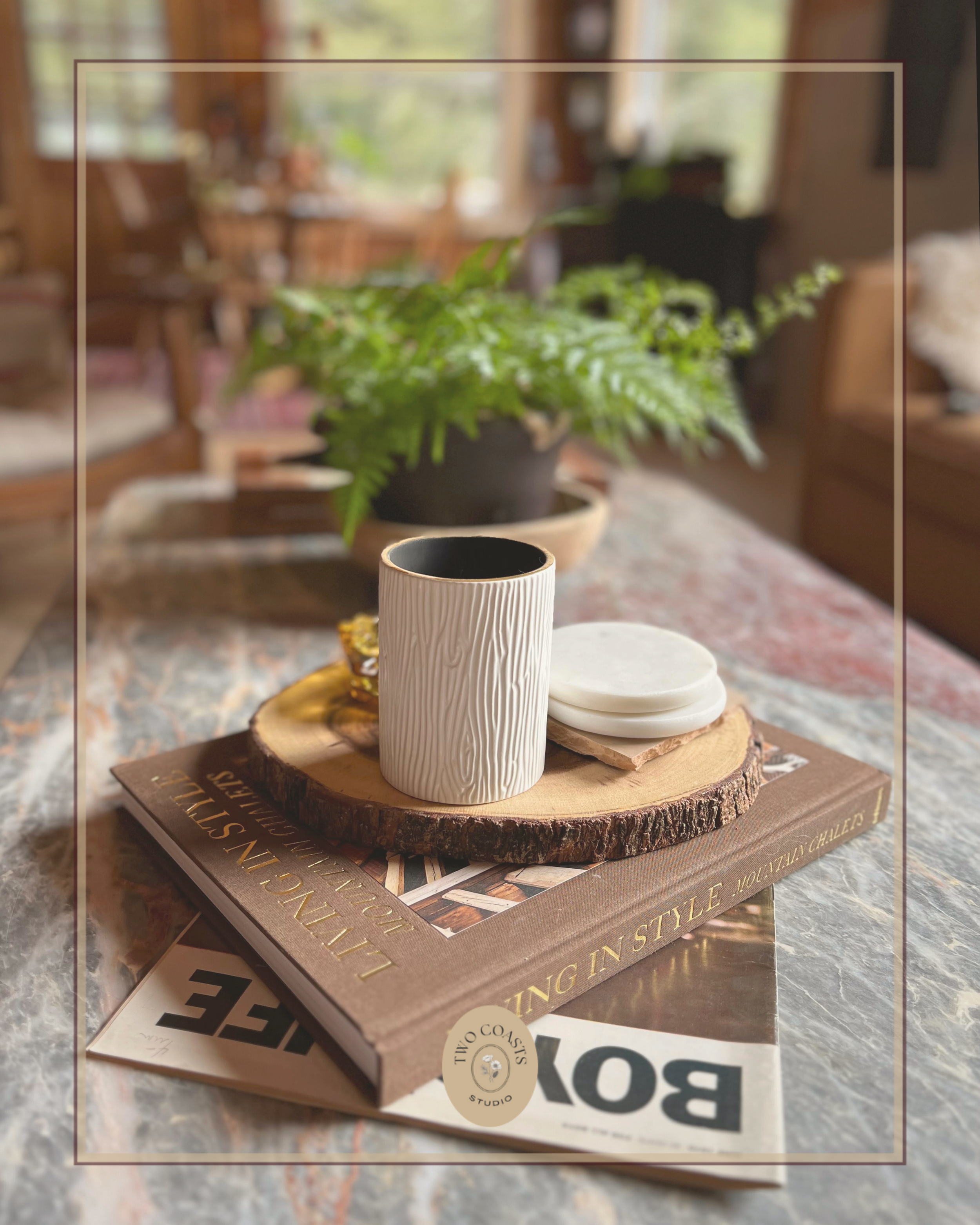 Table with a book, a ceramic mug, a couple of coasters, placed on a round wooden slab, with greenery in the background, in a cozy room.