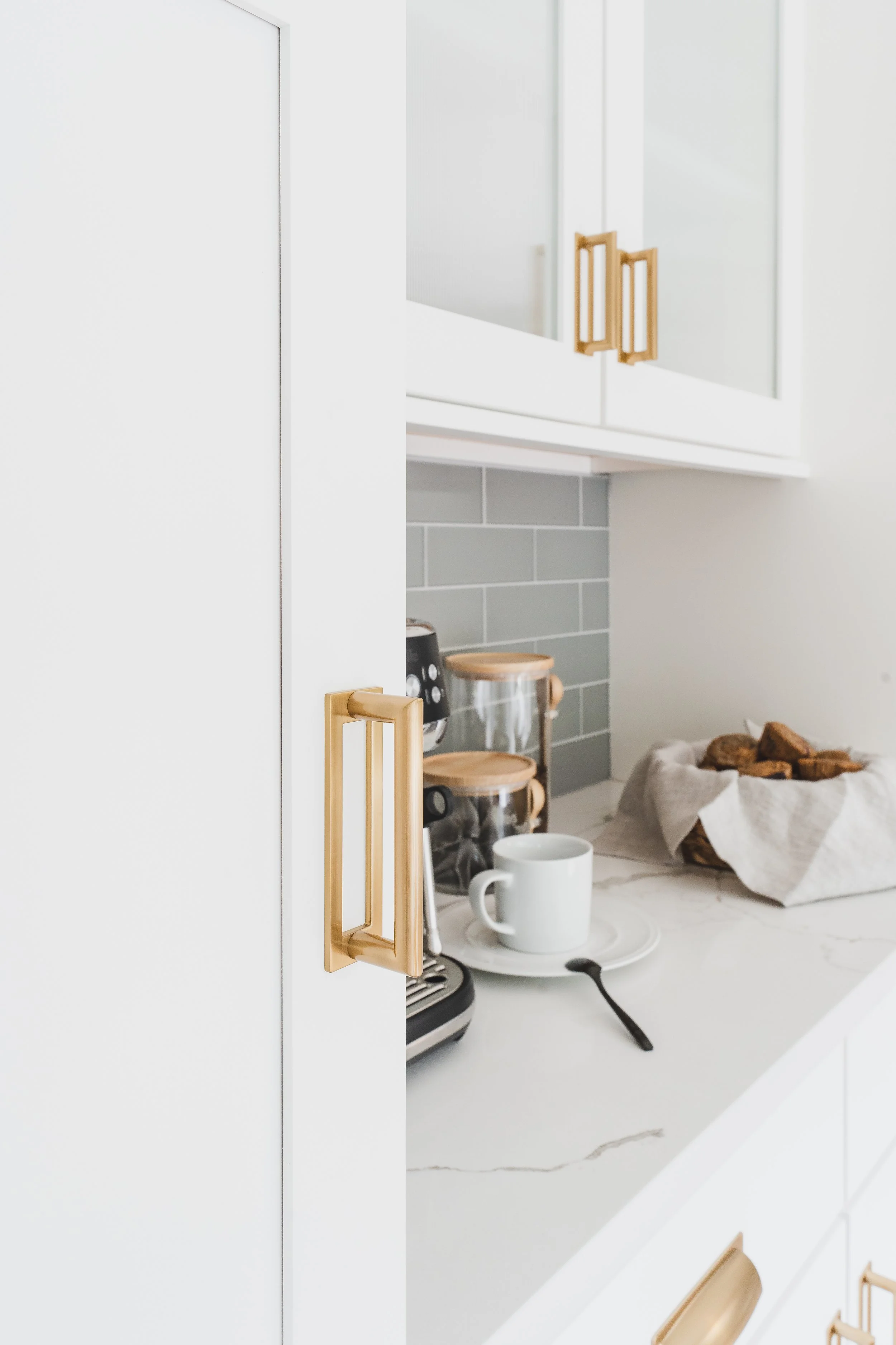 Part of a modern kitchen with white cabinets with gold handles, a countertop with a white mug, a spoon, and a paper bag filled with snacks or bread, and a backsplash made of grey tiles.