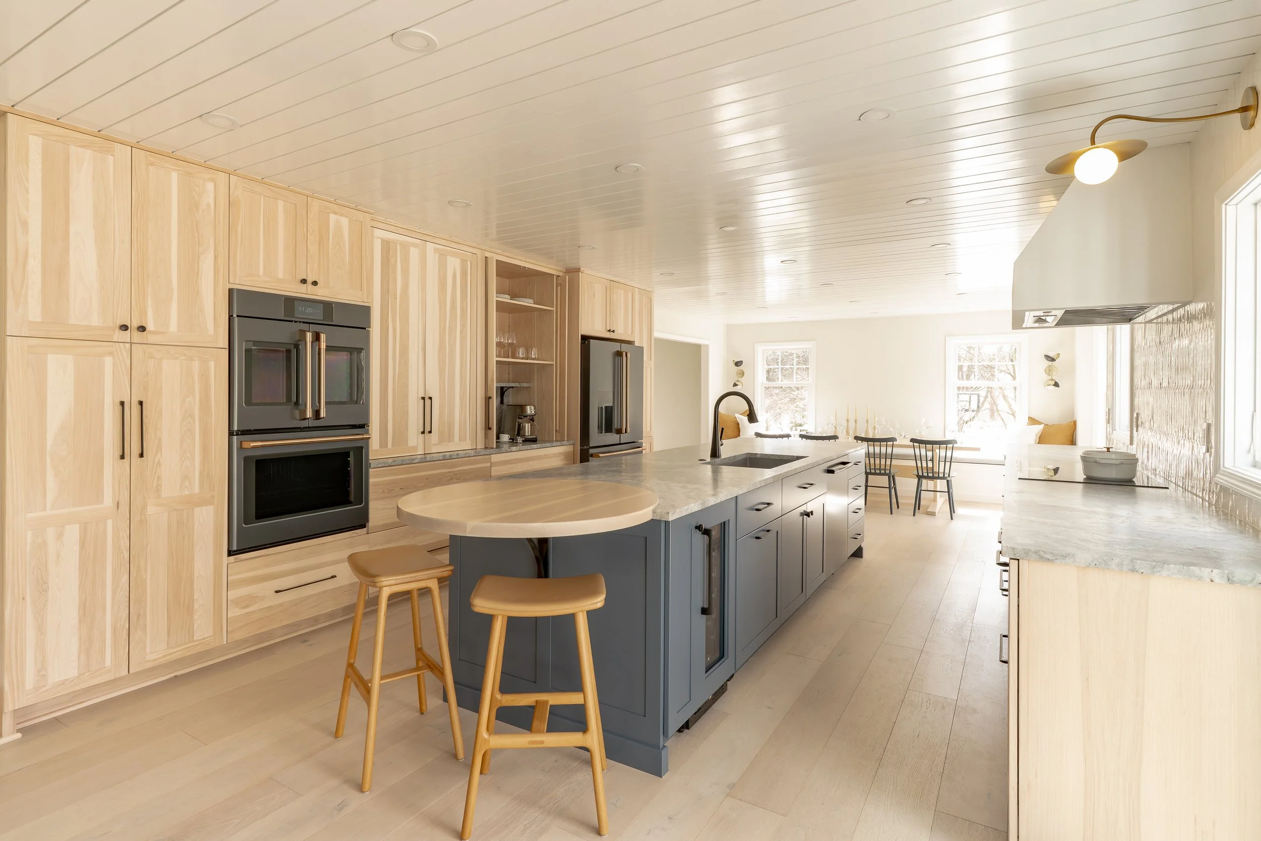 Bright kitchen with light wood cabinets, a blue island with marble countertop, and dining area with windows and seating.