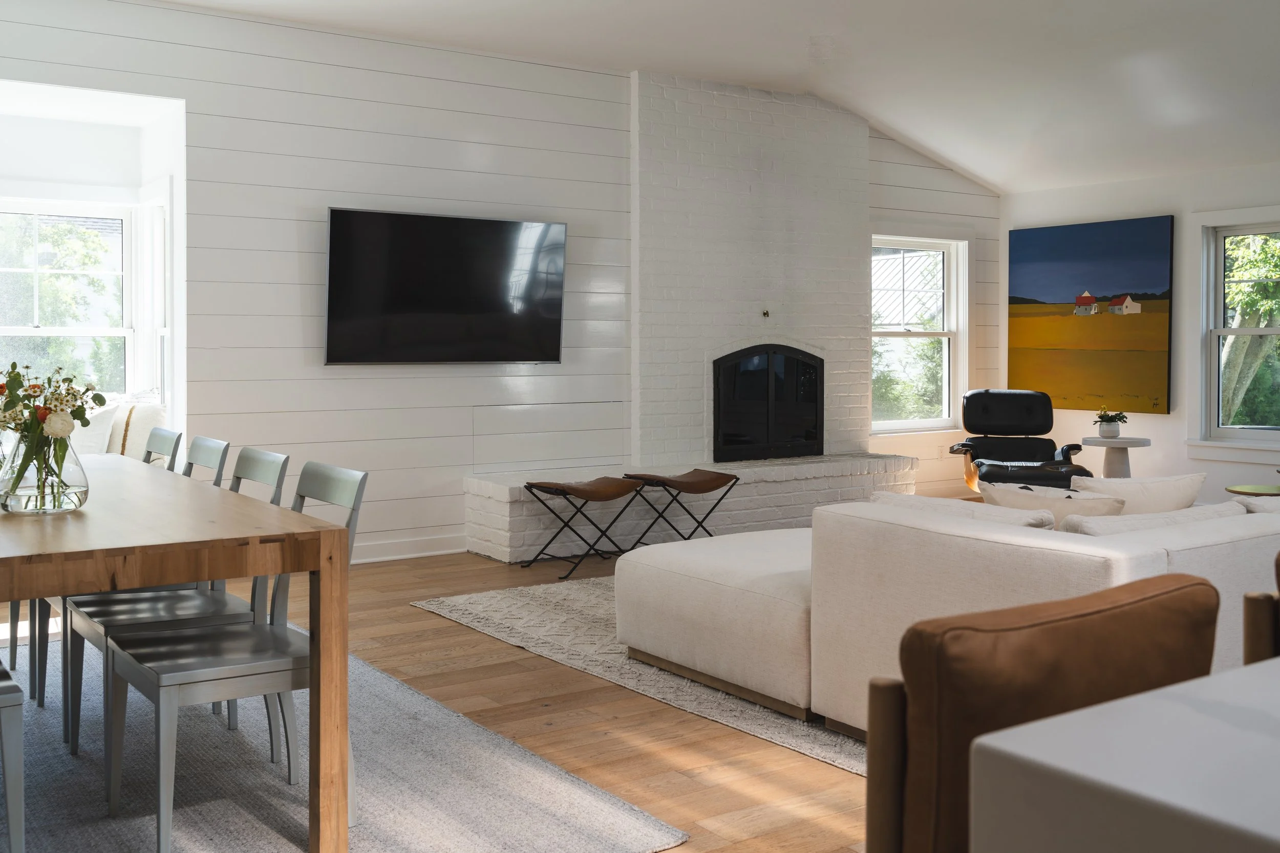 Modern living room with white walls, a wall-mounted flat-screen TV, white brick fireplace, and large windows with views of greenery. The room features a white sofa, a black office chair, a wooden dining table with chairs, and artwork on the walls.