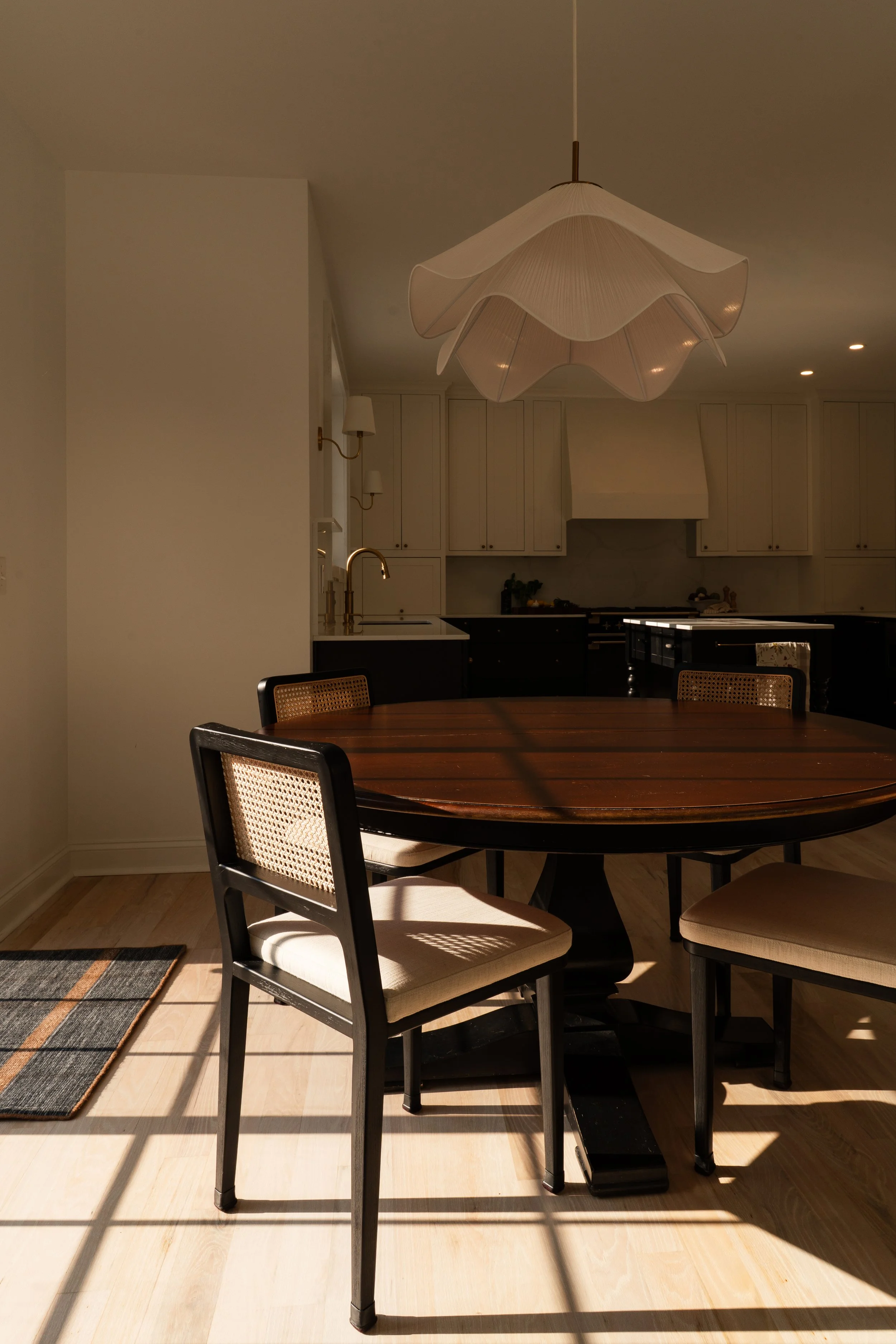 Empty dining room with a round wooden table, four chairs, a hanging lamp, and a kitchen in the background with white cabinets.