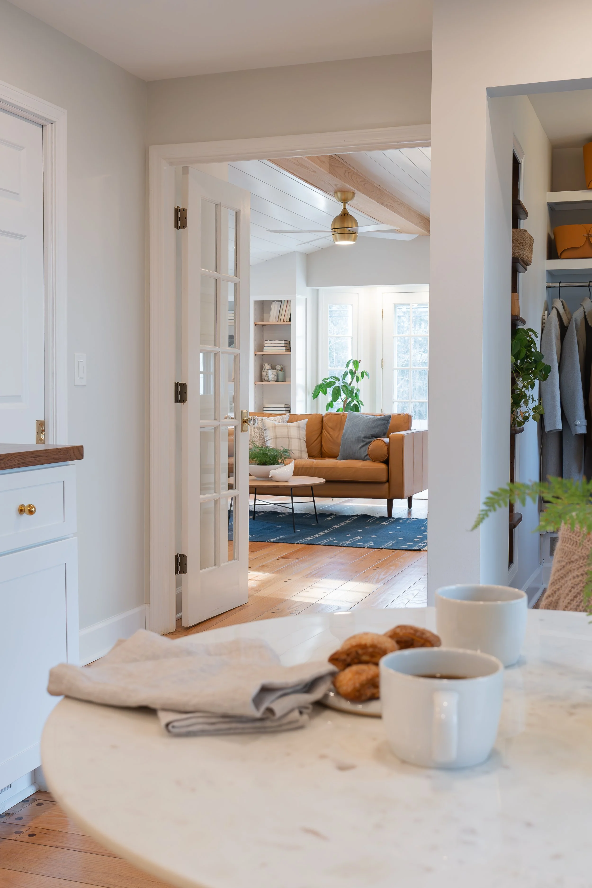 View of a cozy living room with sunlight streaming through large windows, featuring a tan sofa, a round coffee table, and houseplants, seen through a doorway from a kitchen area with two cups of coffee and cookies on the counter.