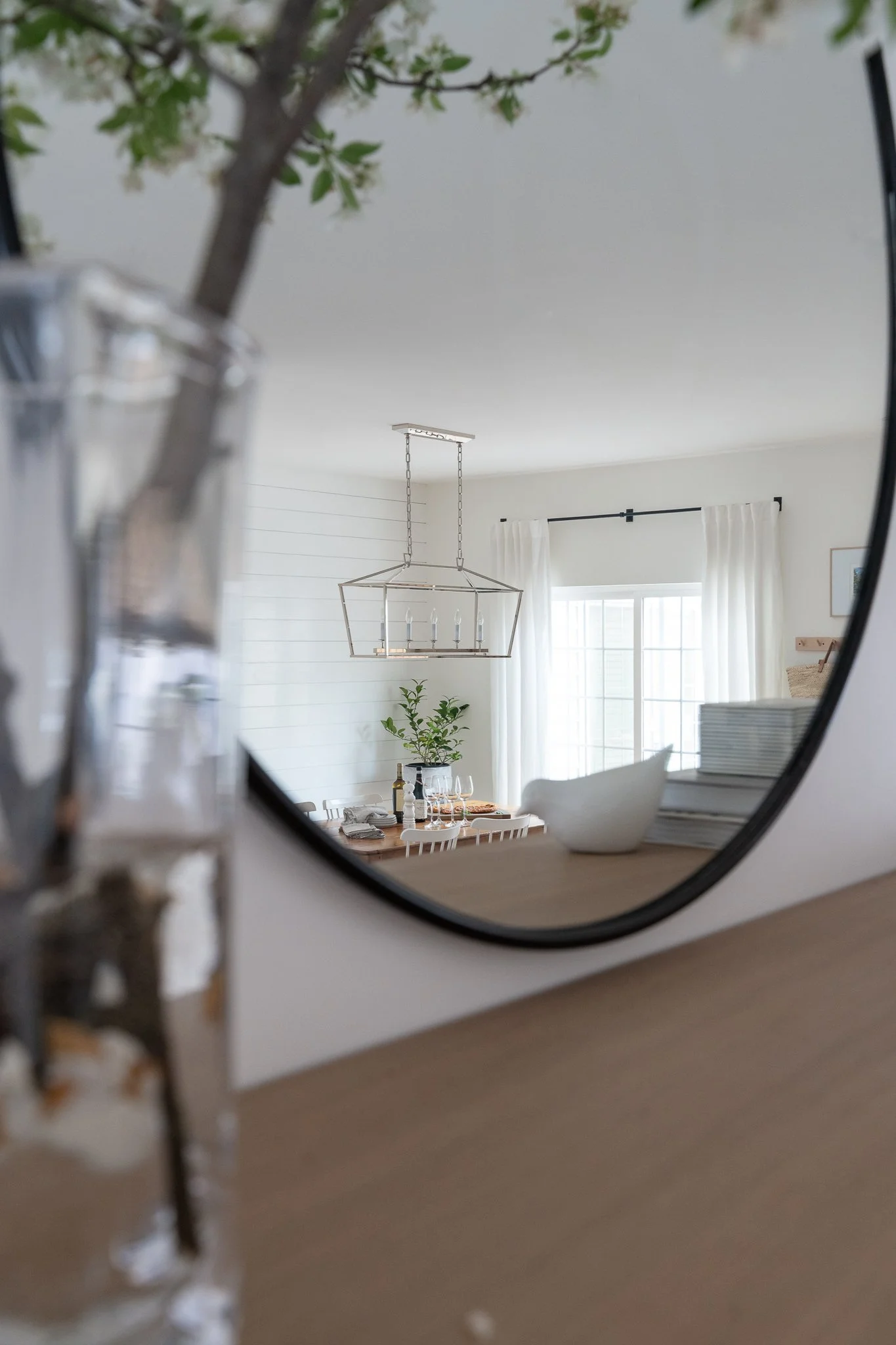 Reflection in a mirror showing a dining room with a chandelier, a window with white curtains, a table set with glasses, a plant, and a stack of plates.