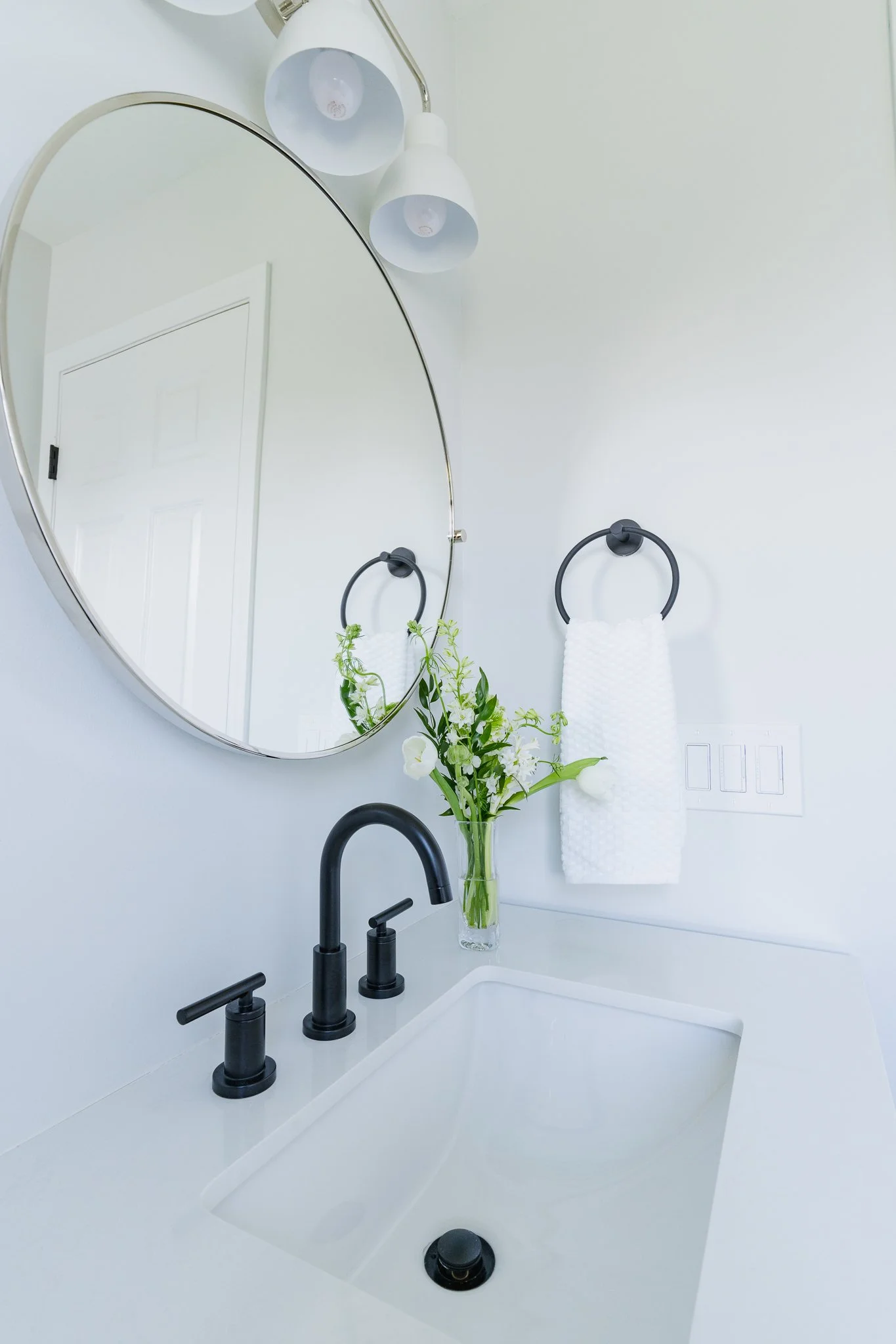 A modern bathroom with a large round mirror above a white sink with black fixtures. There is a vase with flowers on the sink and two white towels on black towel rings. White walls and a white door are visible in the background.
