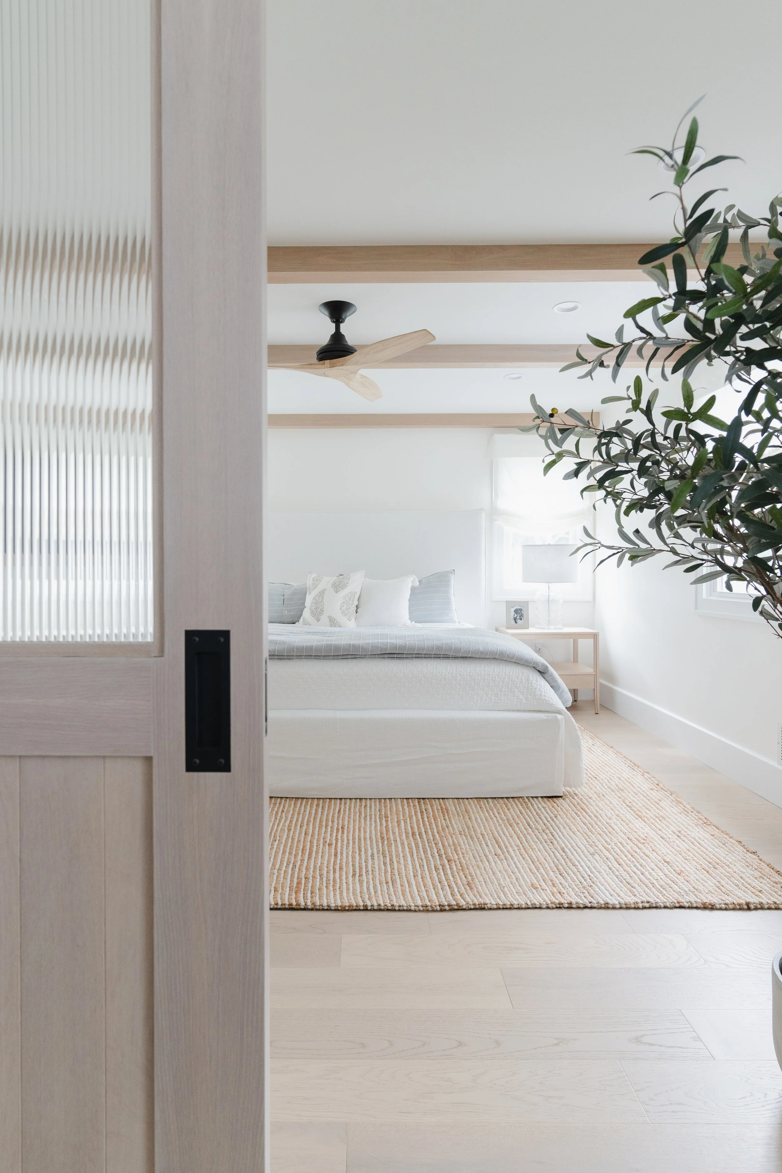 A minimalist bedroom with a white bed, light wood flooring, a beige rug, a nightstand with a lamp, and a window with white curtains, seen through an open sliding door.