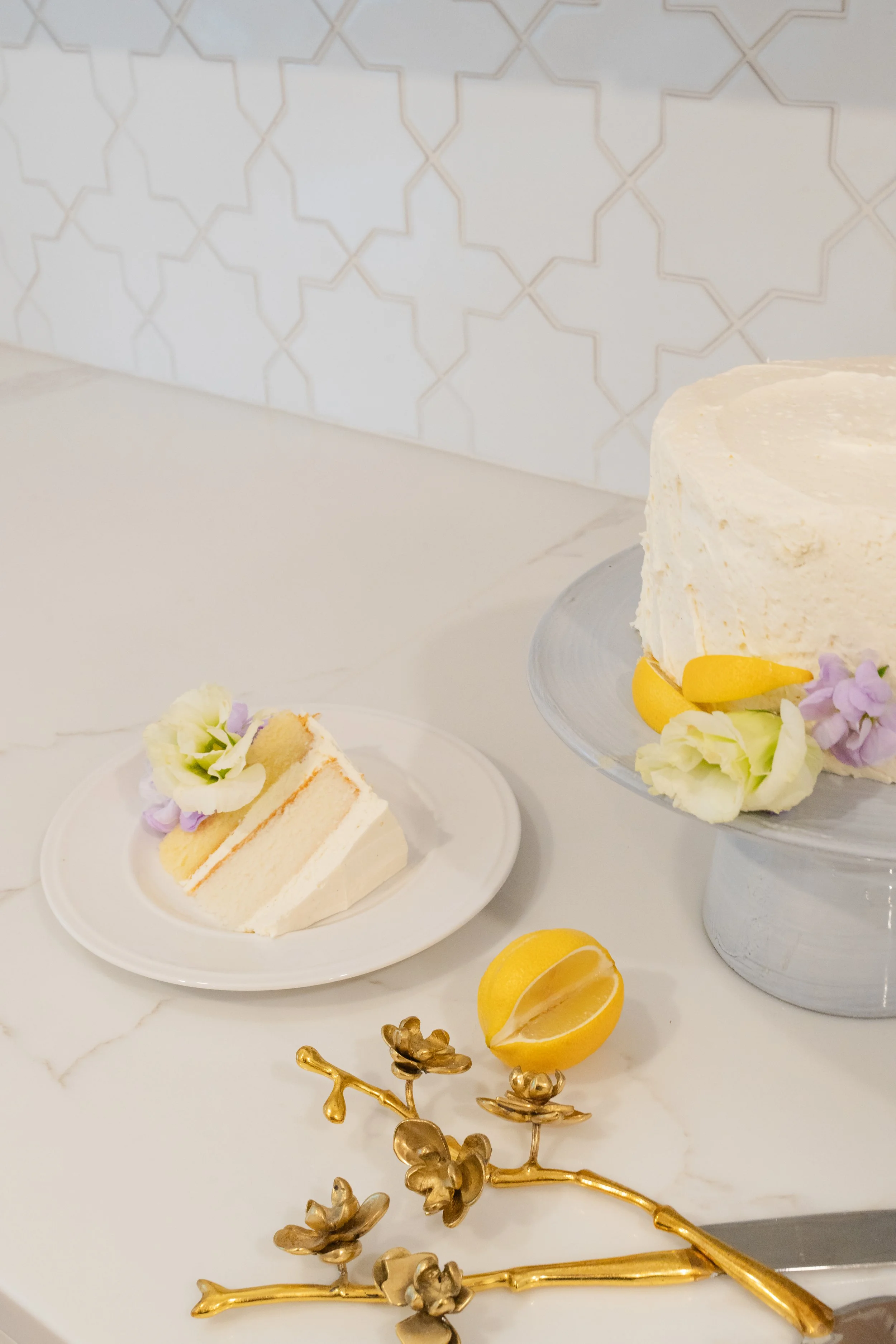 A slice of birthday cake with floral decorations on a small plate, a whole cake decorated similarly on a stand, a halved lemon, and a decorative golden floral ornament on a white marble surface.