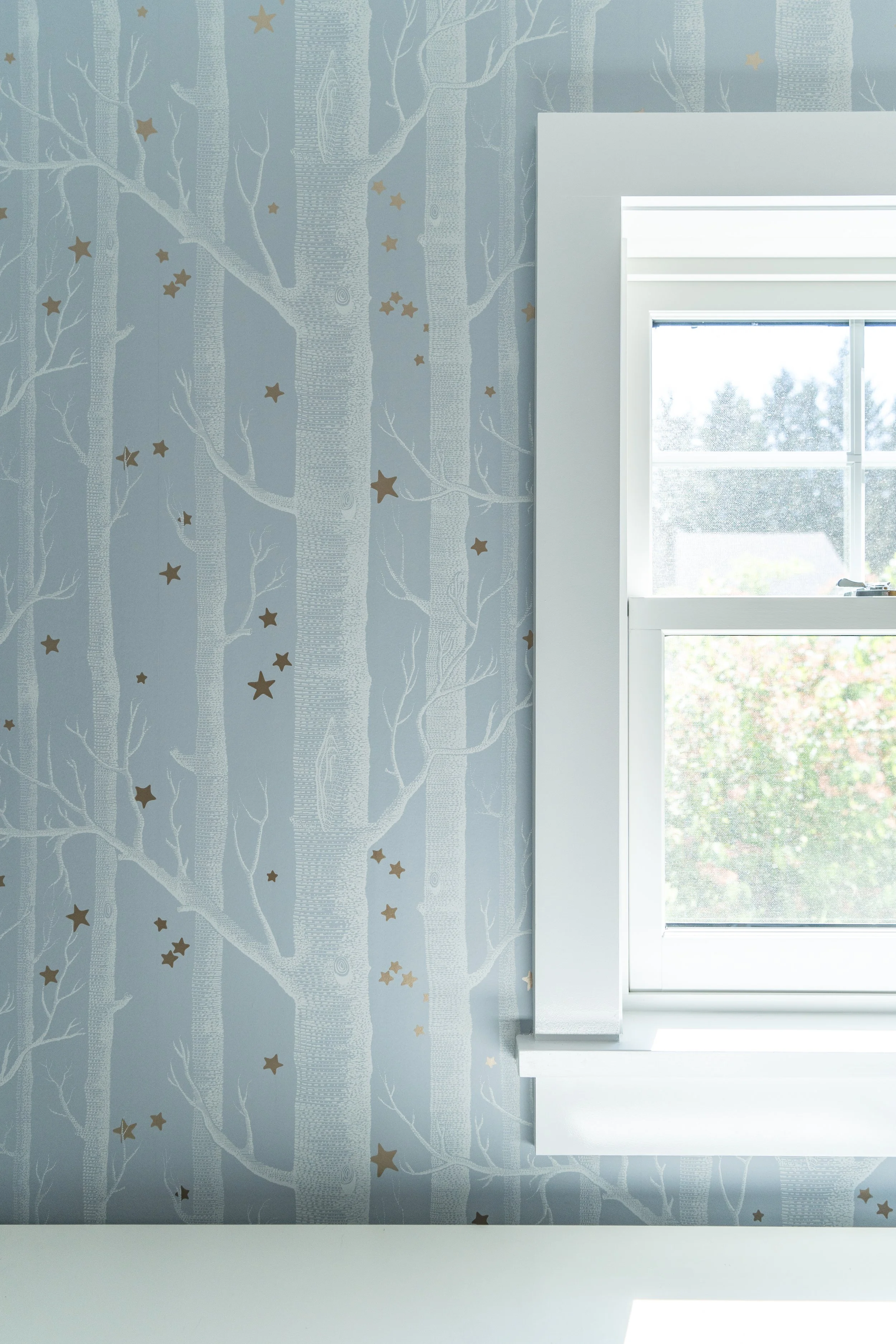 Close-up of a wall with blue wallpaper featuring white tree silhouettes and gold stars, next to a bright window with white framing.