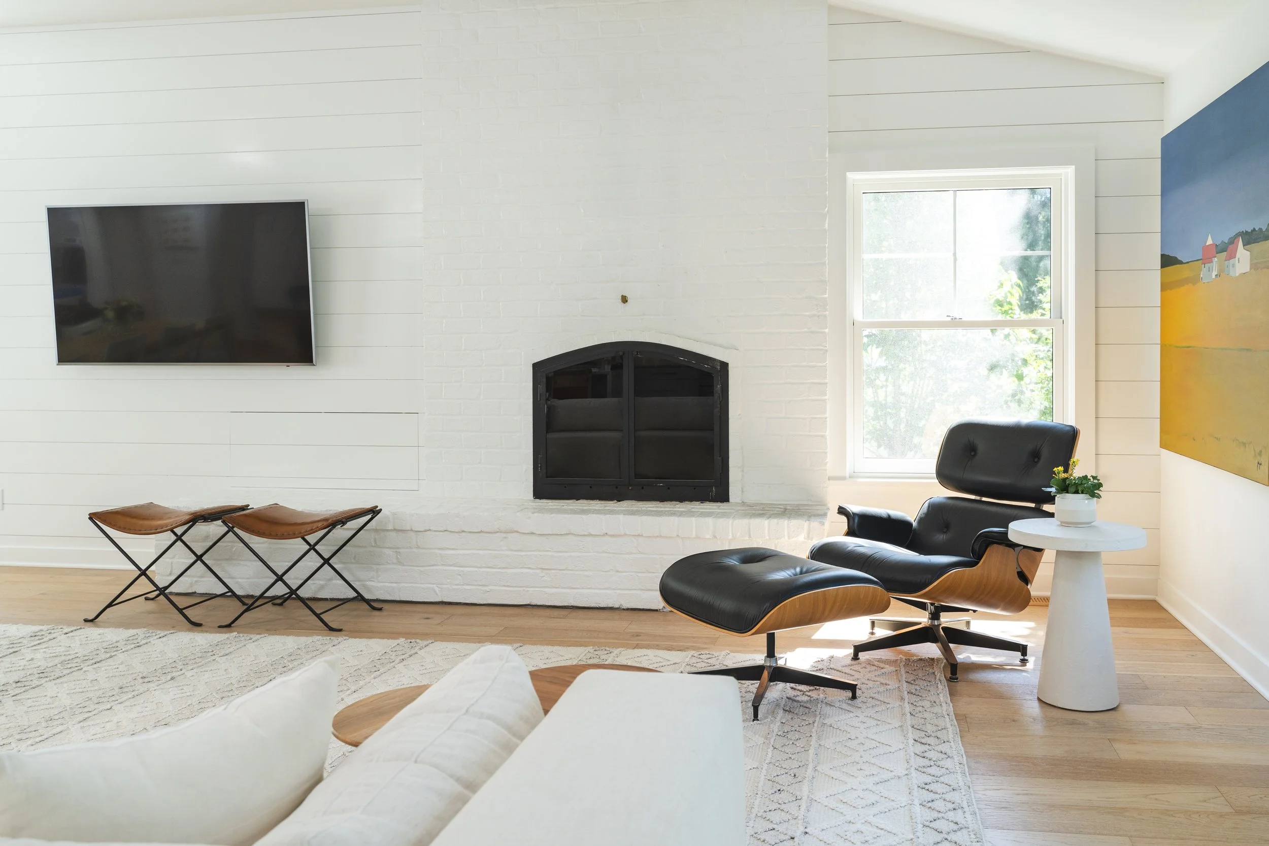 Living room with white brick fireplace, black chair with wooden base, side table with potted plant, large window, mounted TV, and colorful landscape painting