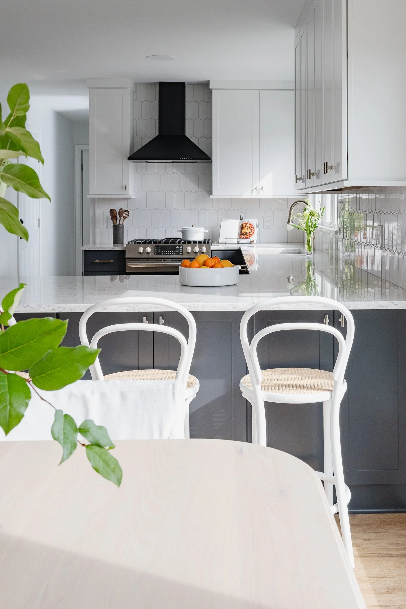 Modern kitchen with white cabinetry, a black range hood, a marble countertop, a bowl of oranges, and white chairs around a light-colored table.