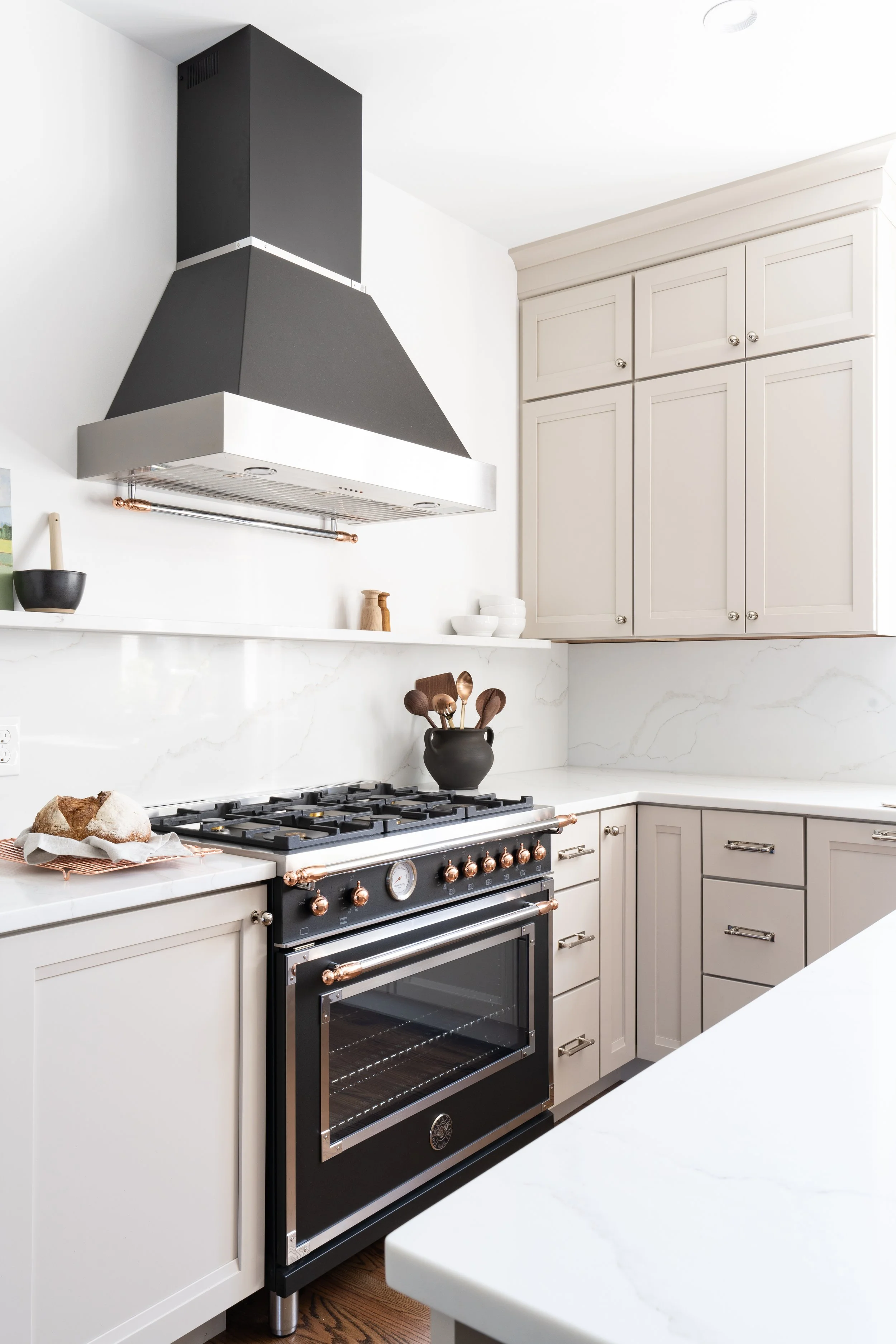 Modern kitchen with white cabinets, marble countertops, and a black and copper range stove. A black pot with wooden spoons is on the stove, and a loaf of bread is on the counter nearby.