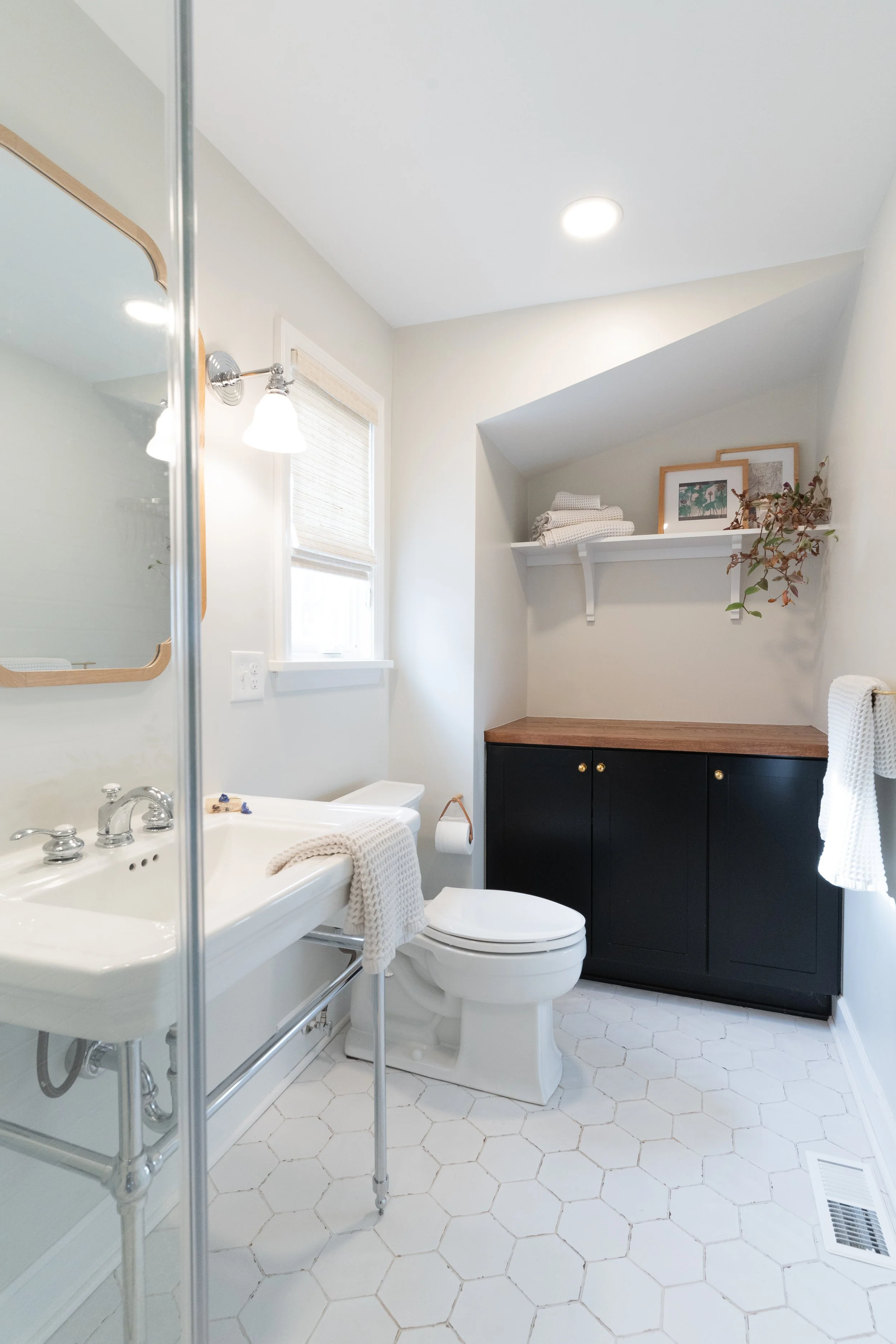 A clean, minimal bathroom with white hexagonal tile flooring, a white pedestal sink, a toilet, a window with a bamboo shade, a black cabinet with a wooden countertop, and a shelf with folded towels and framed artwork.