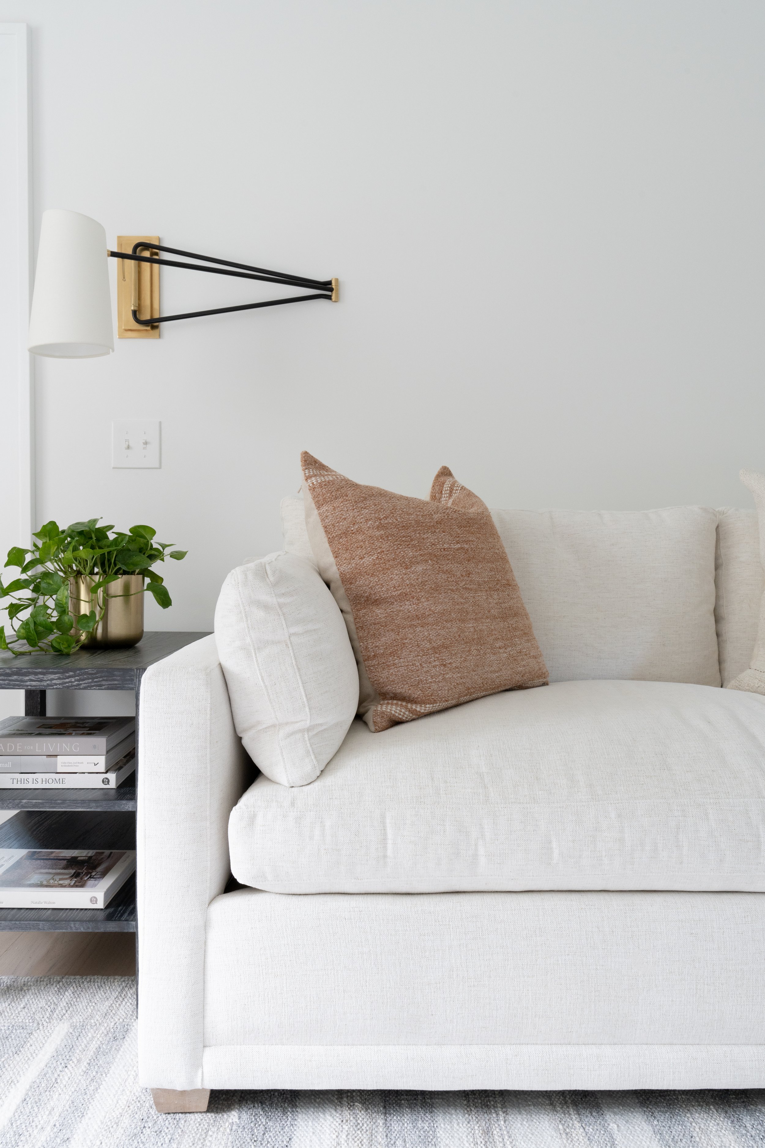 A living room with a white sofa, beige pillows, a black side table with books and a small green plant, and a wall-mounted lamp with a white shade and brass accents.