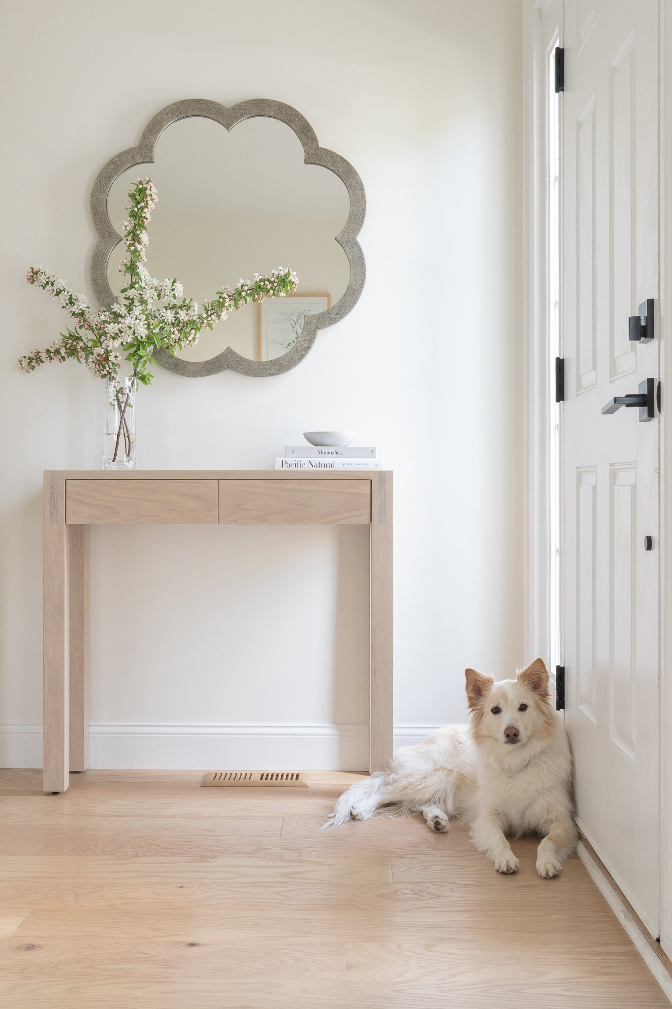 A white dog with fluffy fur, lying on the wooden floor beside an interior white door, in a minimalist home entryway with a light wooden console table, a large floral arrangement, a mirror, and some books.