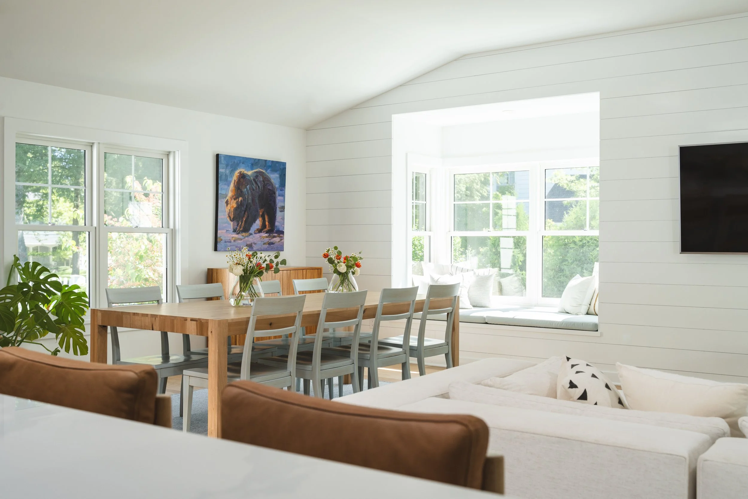 Bright living room with white walls, large windows, a wooden dining table with eight chairs, a window seat with pillows, wall art of a bear, and a television.