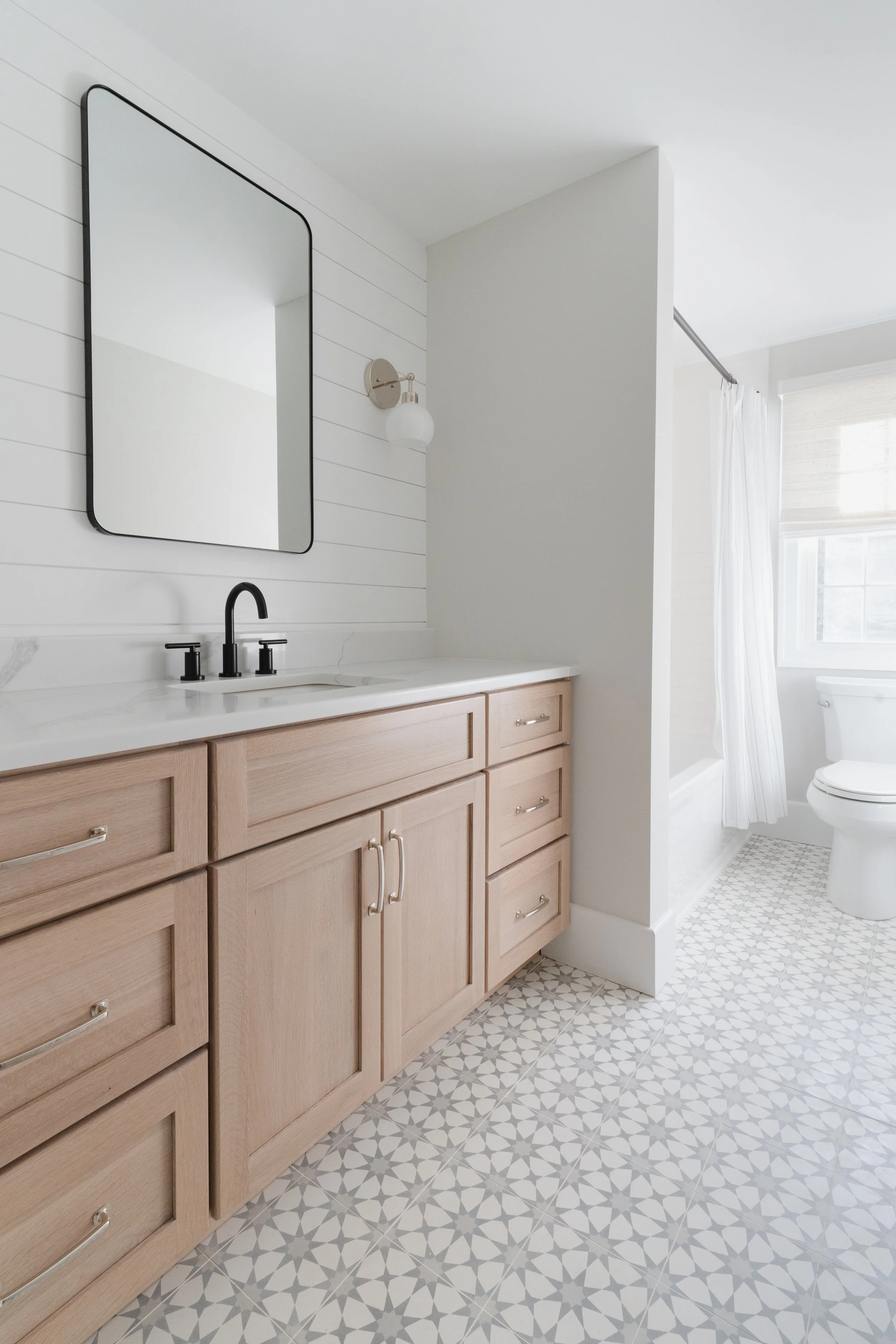 Bathroom with wooden cabinets, marble countertop, rectangular mirror, black faucet, white shiplap wall, wall-mounted light fixture, white toilet, window with blinds, and patterned tile floor.
