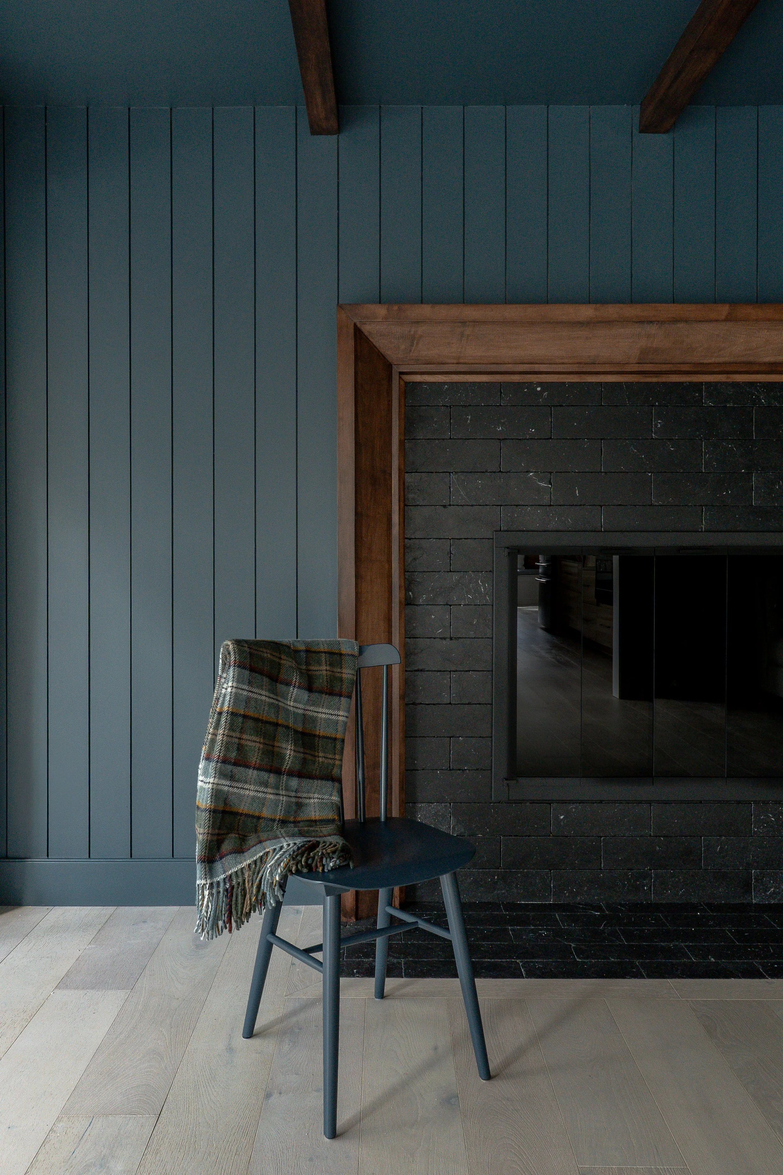 A room with dark blue vertical paneled walls, a black brick fireplace, and a black chair with a tartan blanket draped over it, situated on light-colored wooden flooring.