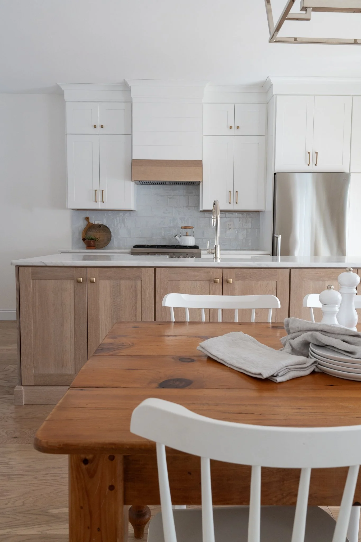 Modern kitchen with white upper cabinets, light wood lower cabinets, a white marble countertop, and a stainless steel refrigerator. A wooden dining table with white chairs and neatly folded napkins.