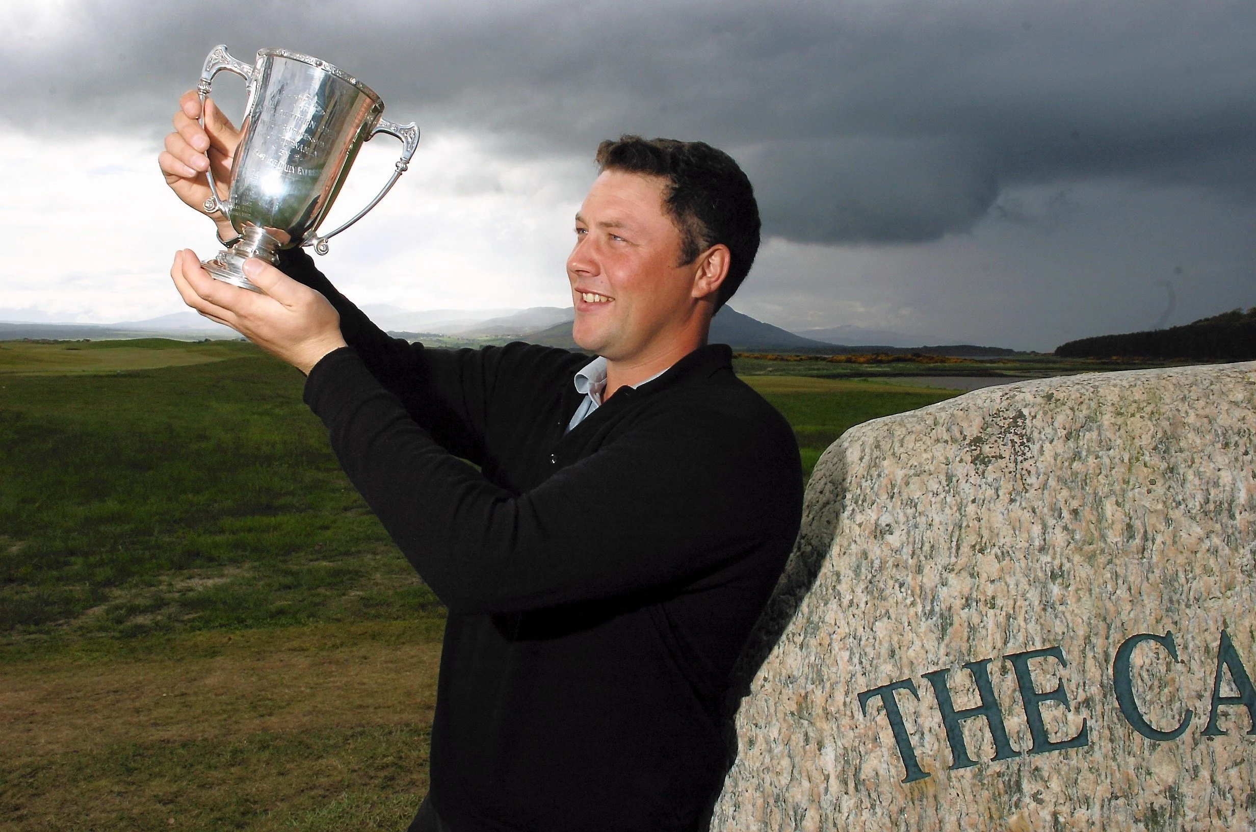 Chris Doak PGA holds a silver trophy up before a cloudy Scottish sky. The Northern Open, Skibo Castle, Scotland in 2005. He smiles before a stone reading The Carnegie Club