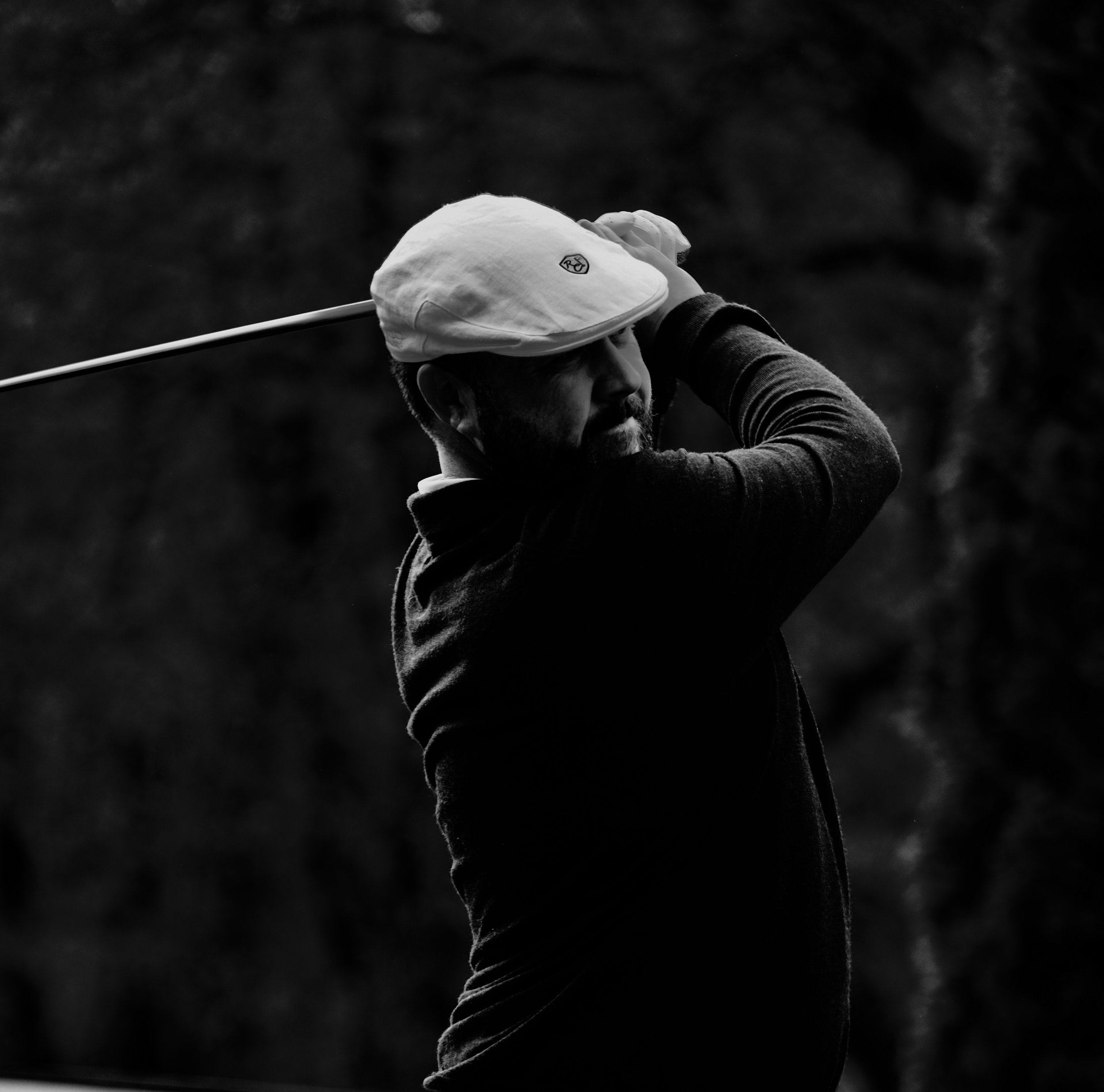 Black and white image of golfer Chris Doak. He wears a white flat cap. He holds the club above his head and looks into the distance after hitting a great golf shot.