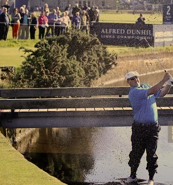 Chris Doak hits his second shot out of a small river. Golf tournament Alfred Dunhill Links Championship St Andrews old Course. He wears a white flat cap. Water and mud splashes across him as he swings