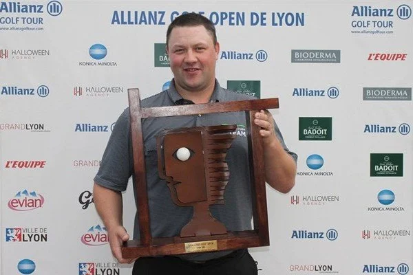 Chris Doak Scottish golfer holds a wooden trophy and poses before sponsorship logos to celebrate his win at the Allianz Golf Open Lyon France in 2012. He smiles. 