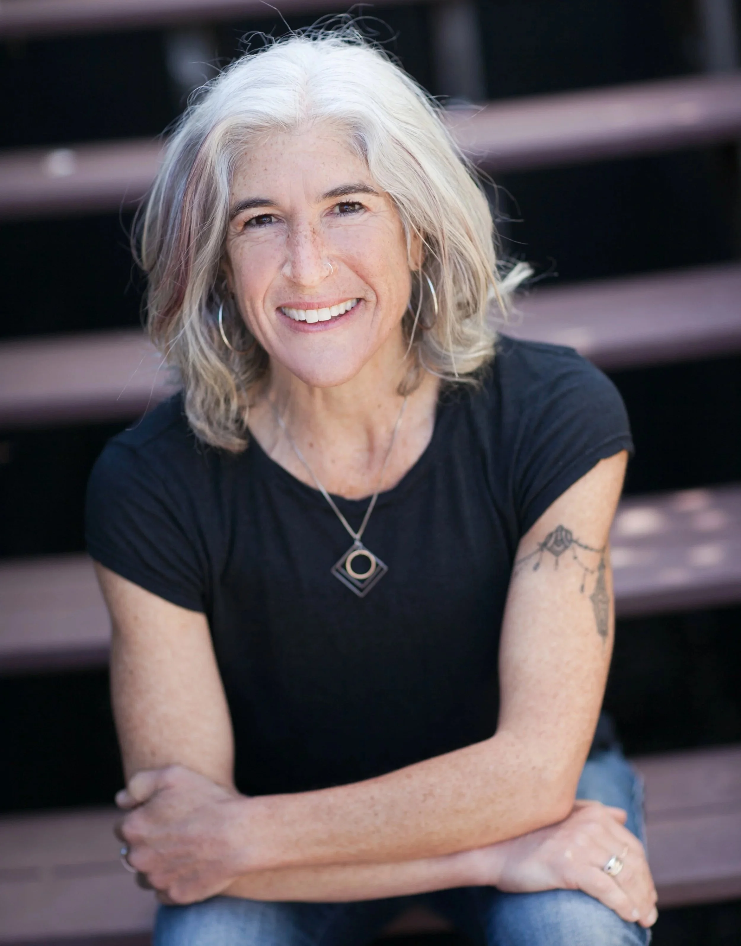 A photo of Rachel Heron; a woman smiling and sitting outdoors on wooden steps. She has grey hair, a nose ring, visible freckles, and a tattoo on her left arm.